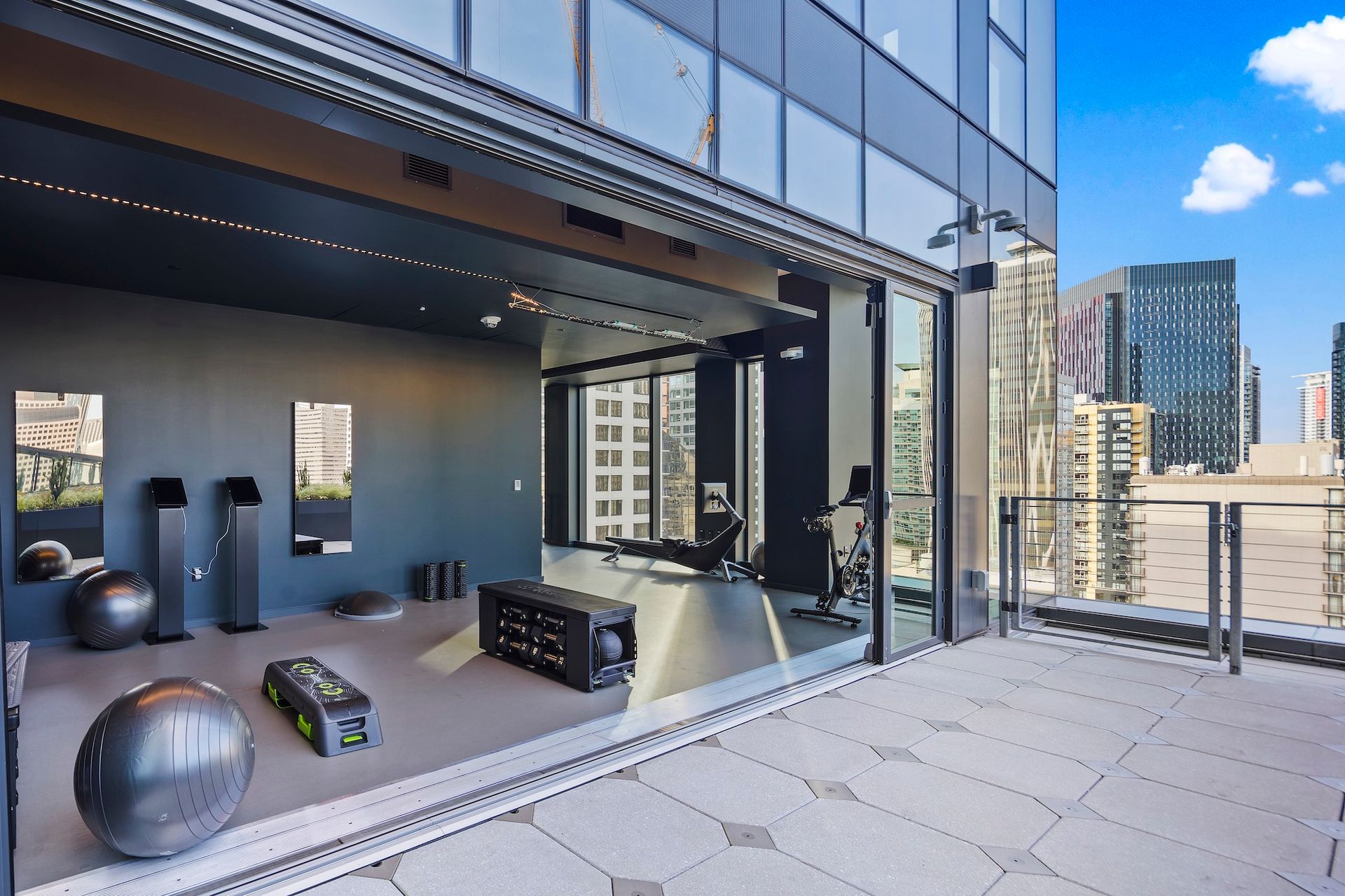 Modern balcony gym with workout equipment, city skyline, and blue sky view.