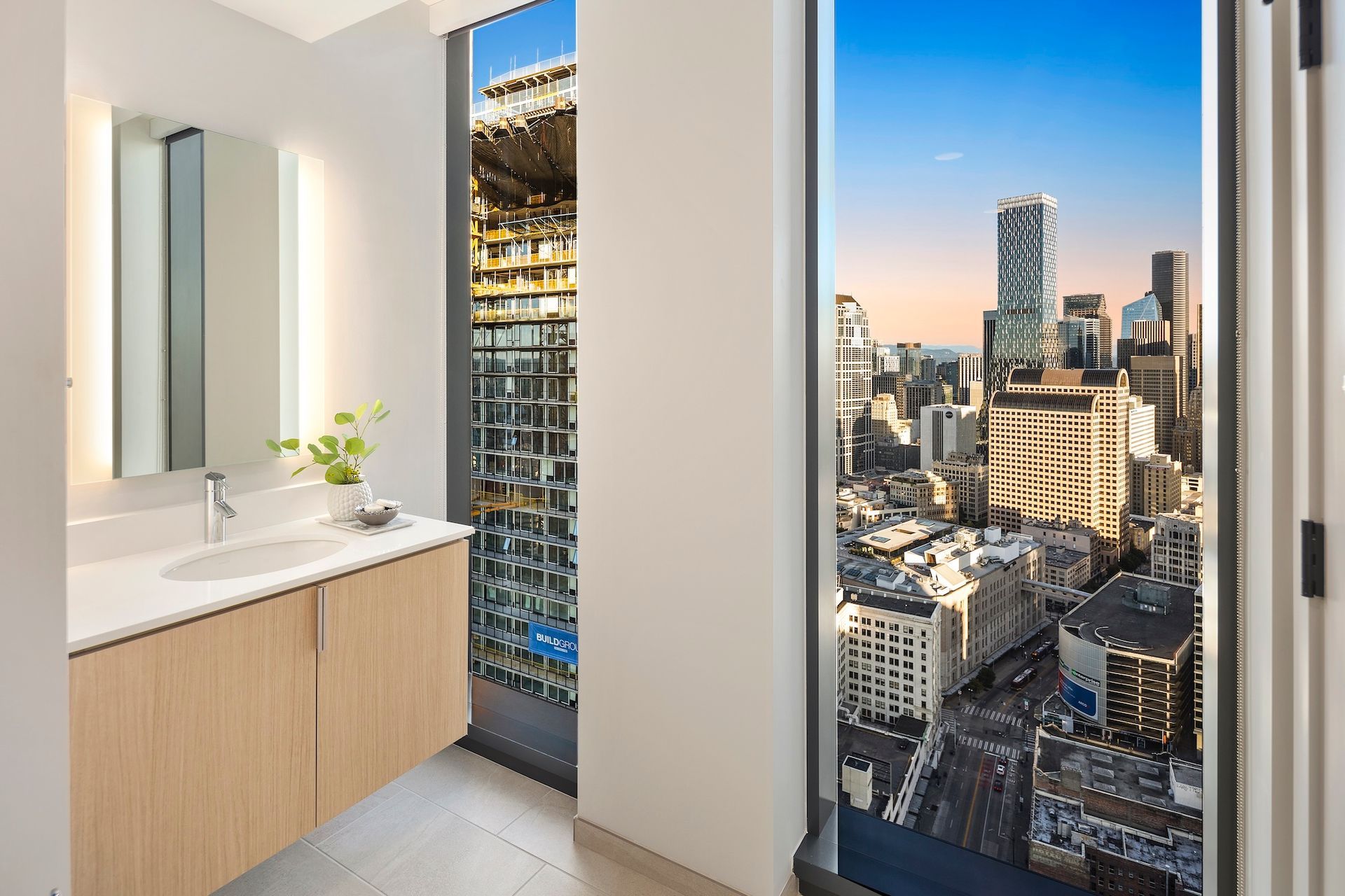 Modern bathroom with vanity and mirror beside floor-to-ceiling windows overlooking a city skyline.