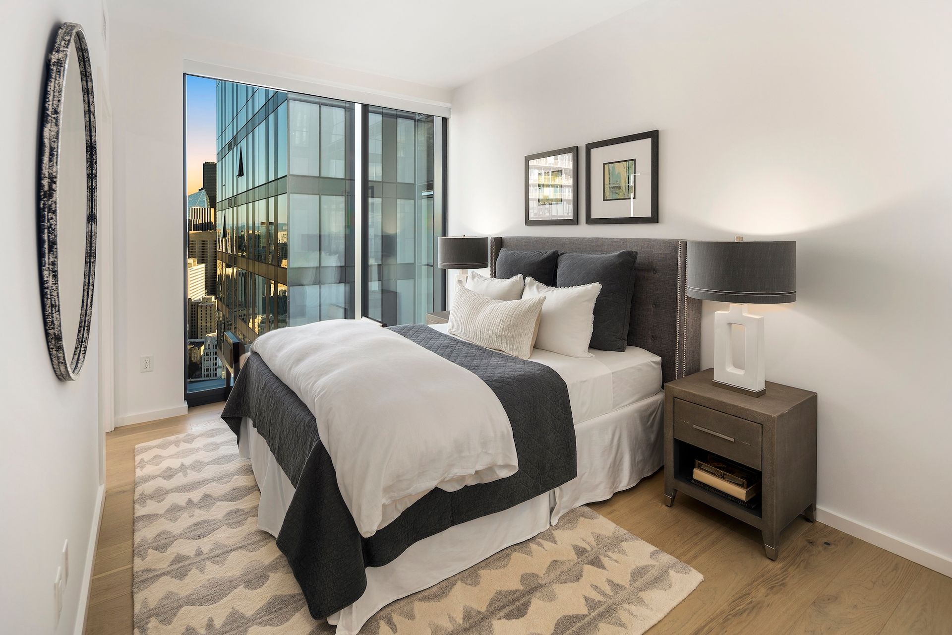 Modern bedroom with a bed, nightstand, framed art, and city view through large windows.