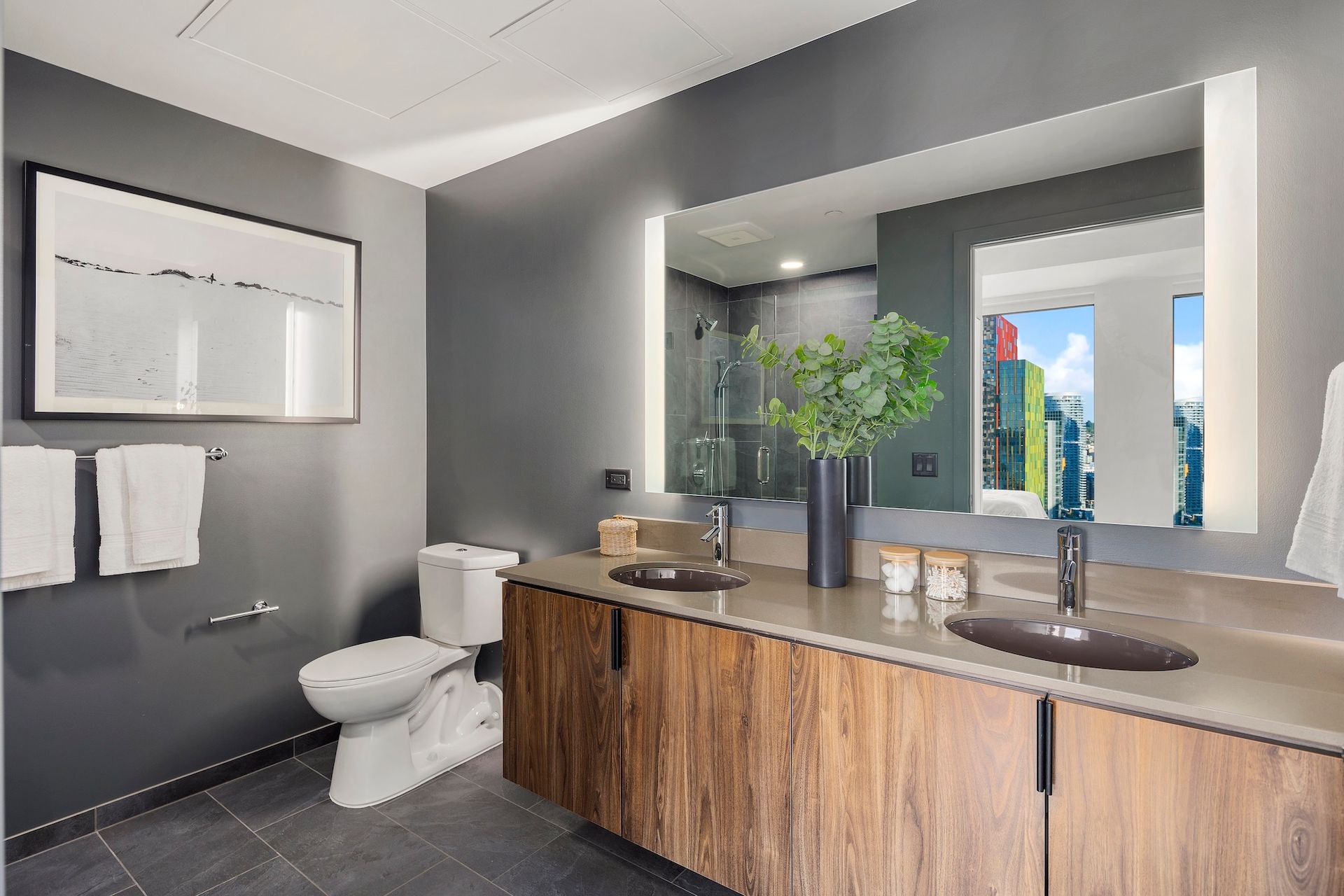 Modern bathroom with wood vanity, large mirror, toilet, and window view of greenery and buildings.