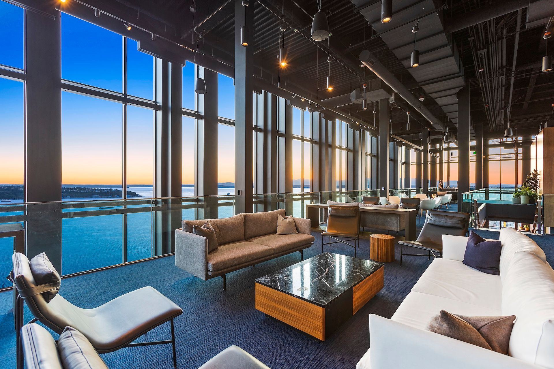 Modern lounge with floor-to-ceiling windows overlooking water at sunset.