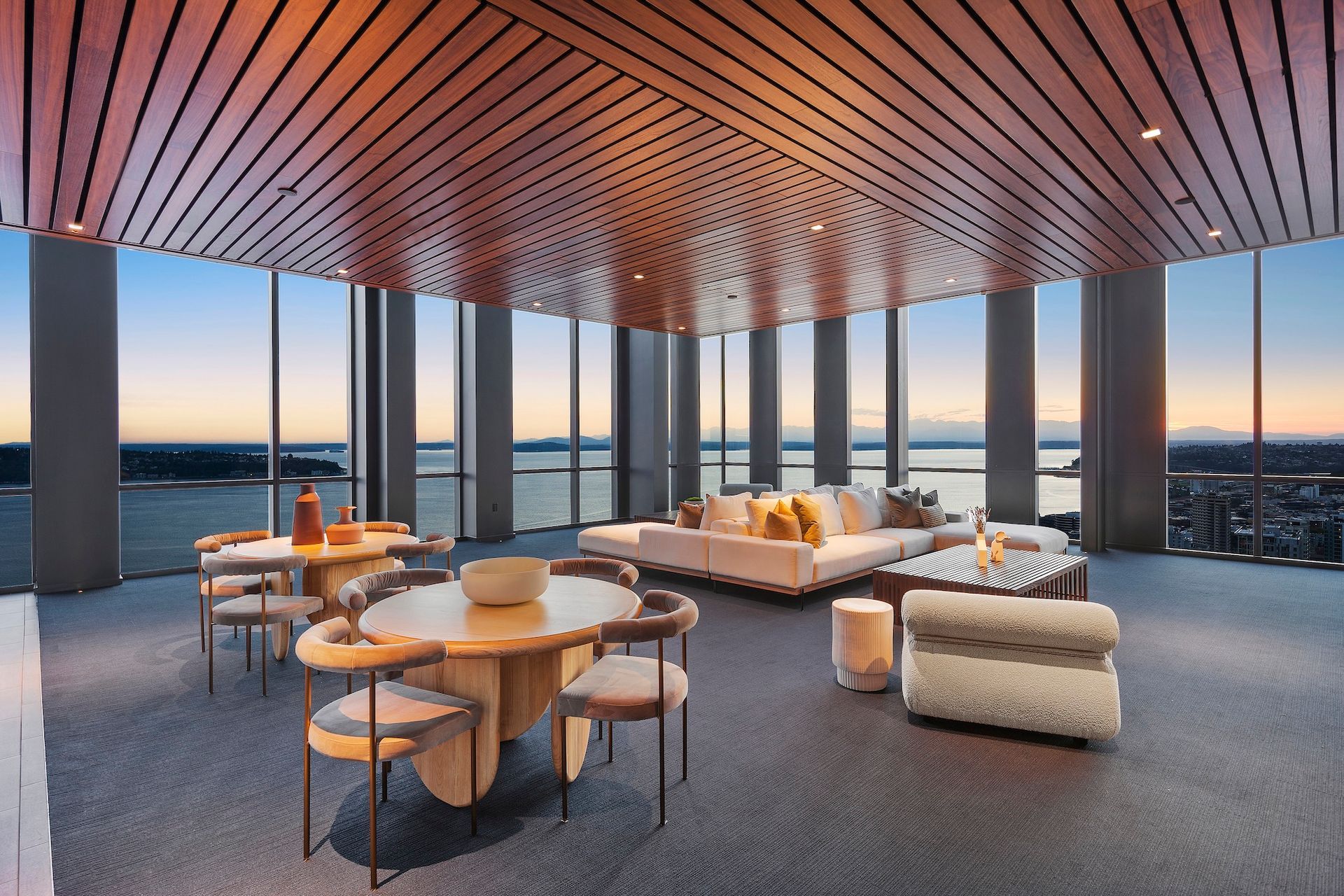 Modern lounge with curved wood ceiling, sofas, and tables overlooking a sunset city skyline.