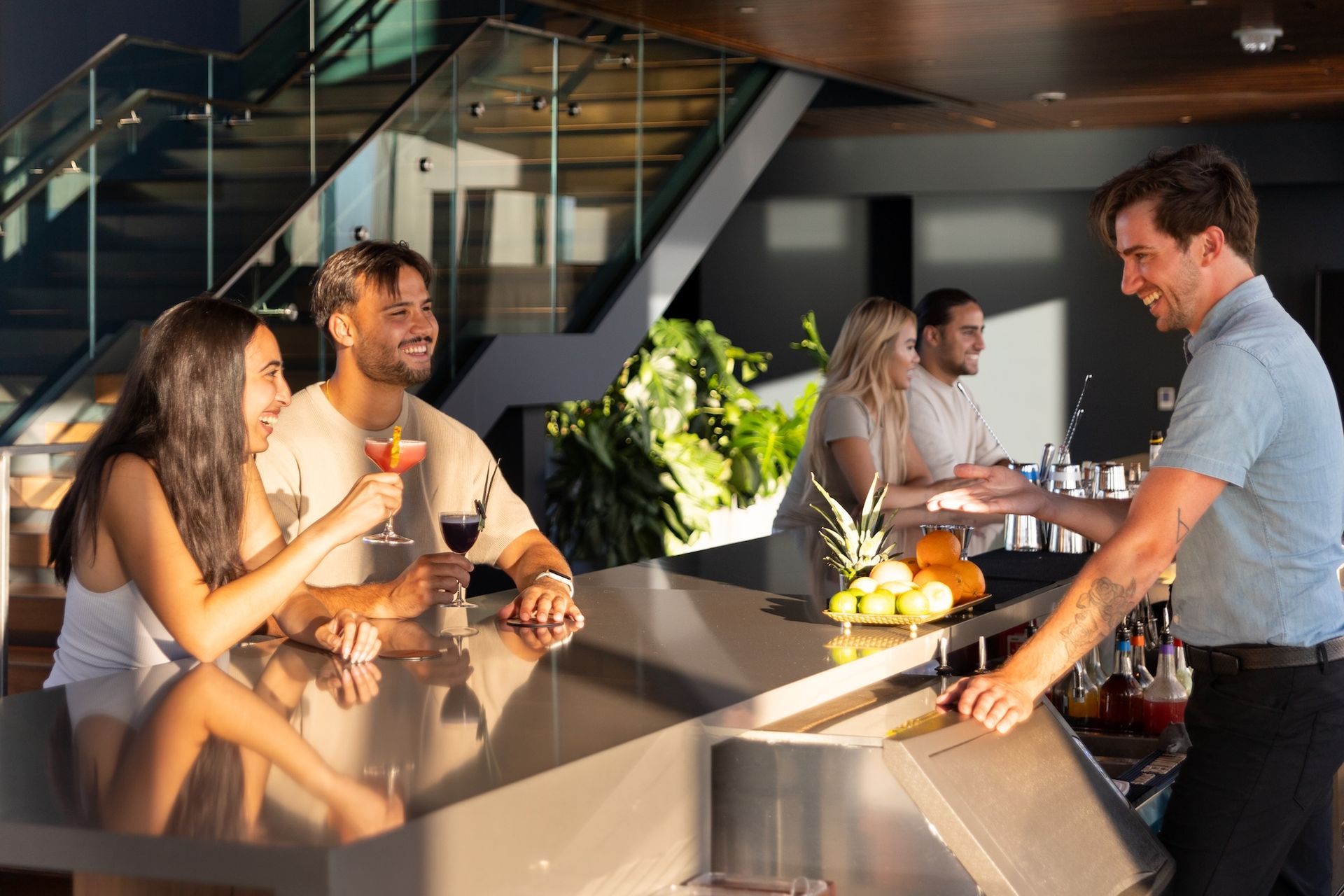 People chatting at a modern bar, holding drinks in warm sunlight.