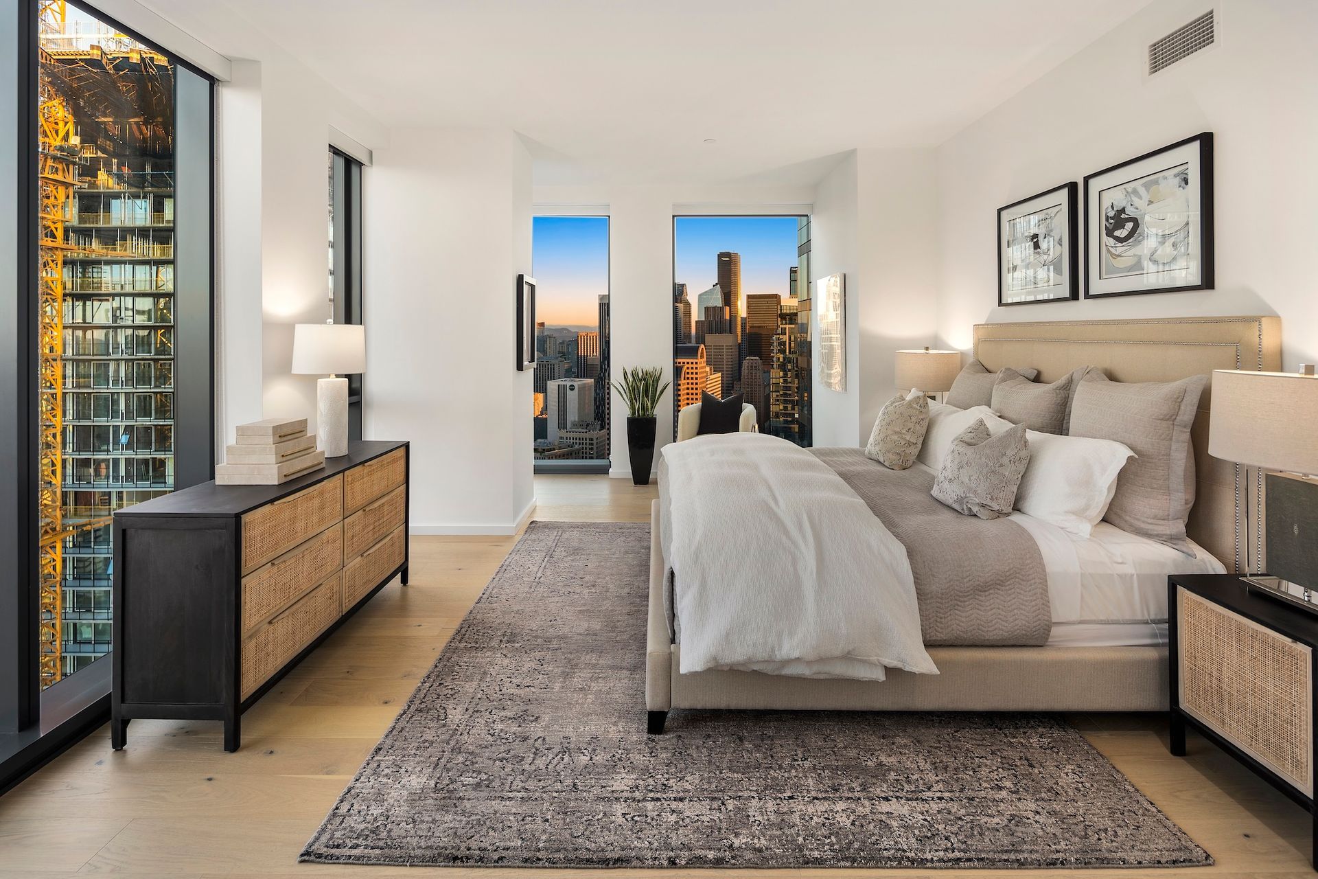 Modern bedroom with a beige bed, gray rug, city skyline view, and black dresser beside floor-to-ceiling windows.