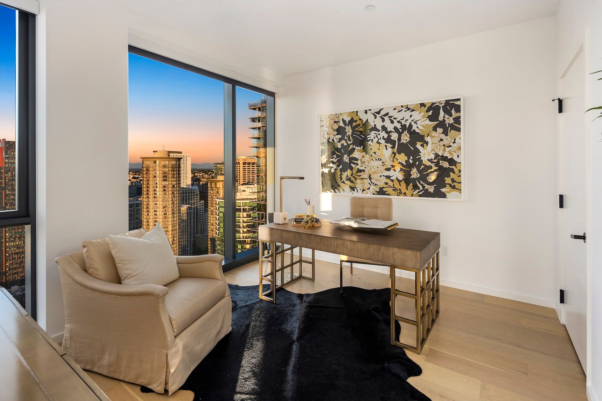 Modern living room with beige armchair, desk, abstract art, and floor-to-ceiling window overlooking city skyline.