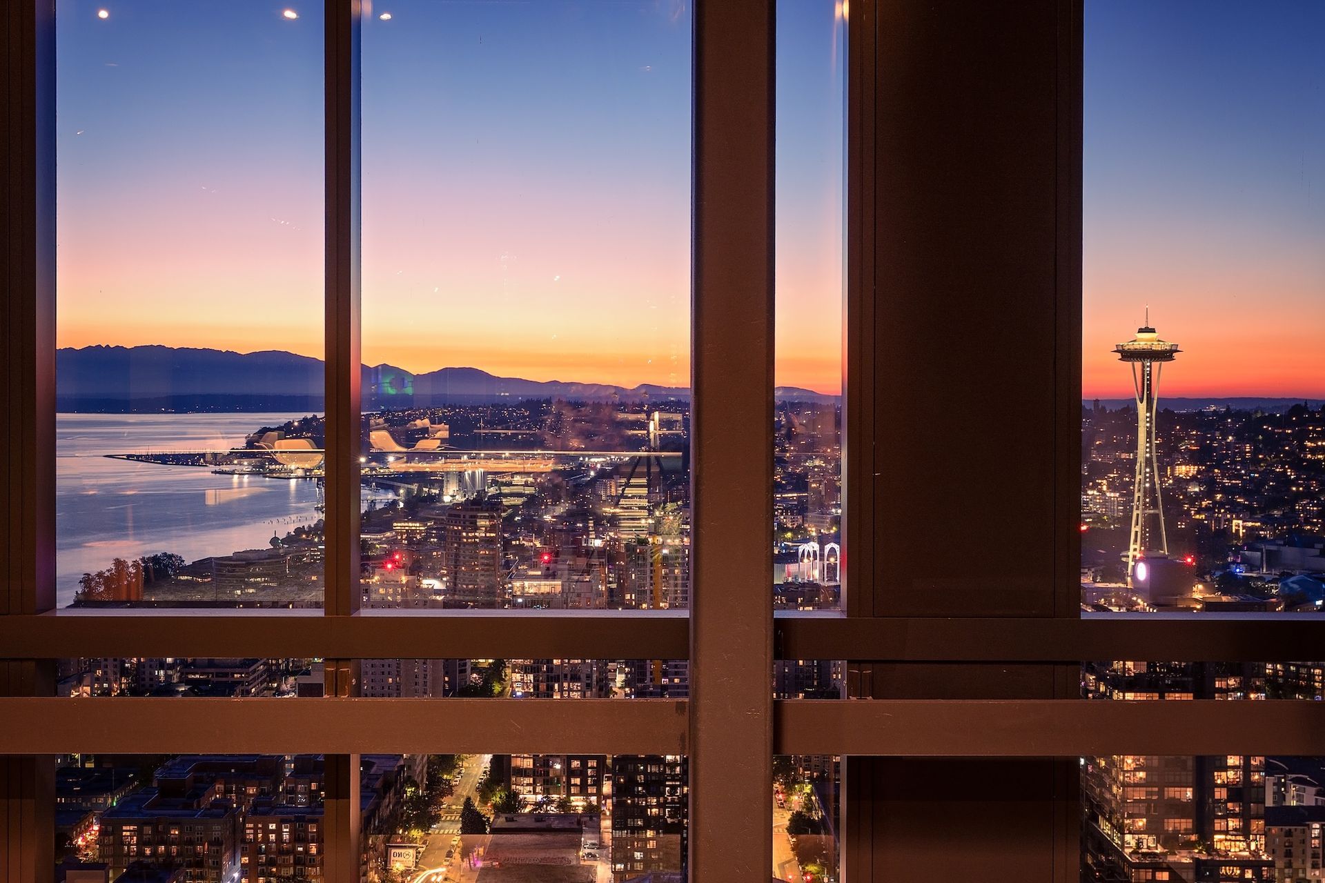 Sunset city skyline seen through a window, with glowing buildings and water in the distance.