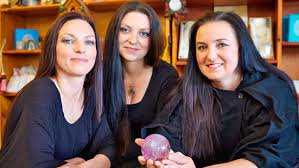 Three people with dark hair and black clothing pose in front of shelves, one holding a spherical pink crystal.