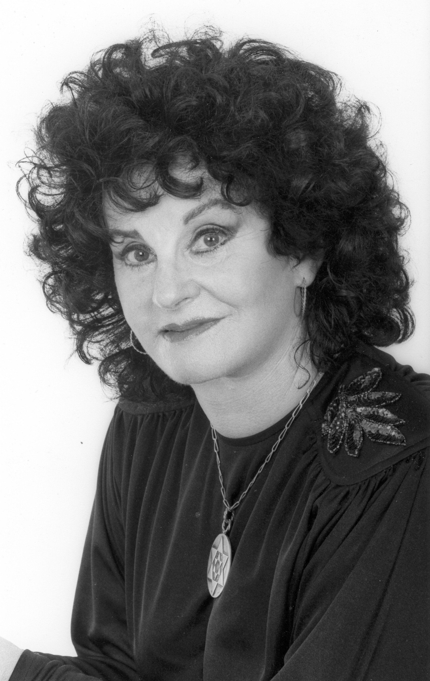 A headshot of a person with dark, curly hair wearing a dark top with a pendant necklace against a white background.