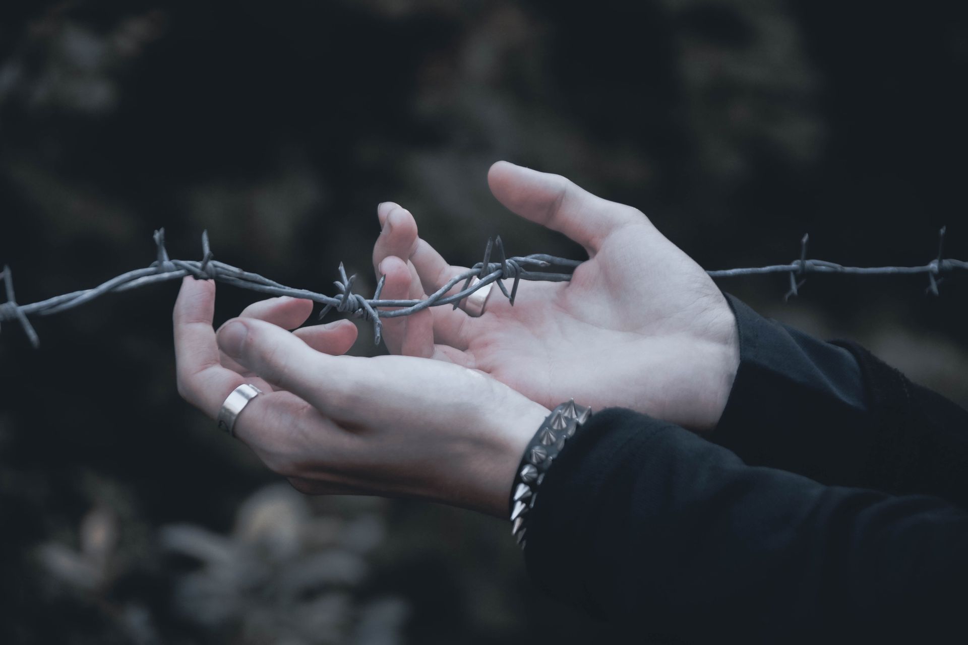 Hands reaching toward barbed wire, one wearing a ring and bracelet, against a dark background