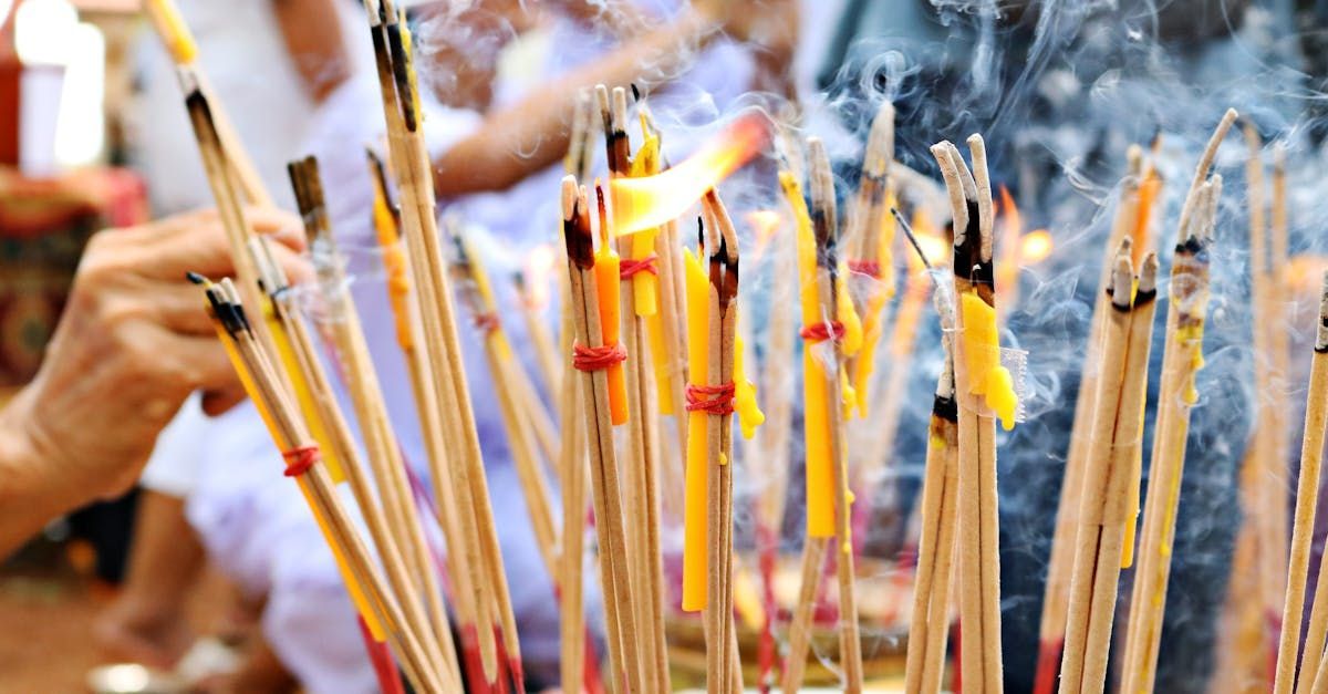 A close-up of a person's hand holding a bundle of burning incense sticks and yellow candles in a smoky temple setting.