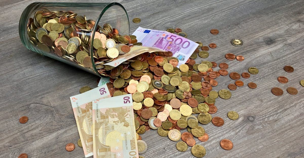 A glass filled with money and coins is spilling out onto a wooden table.