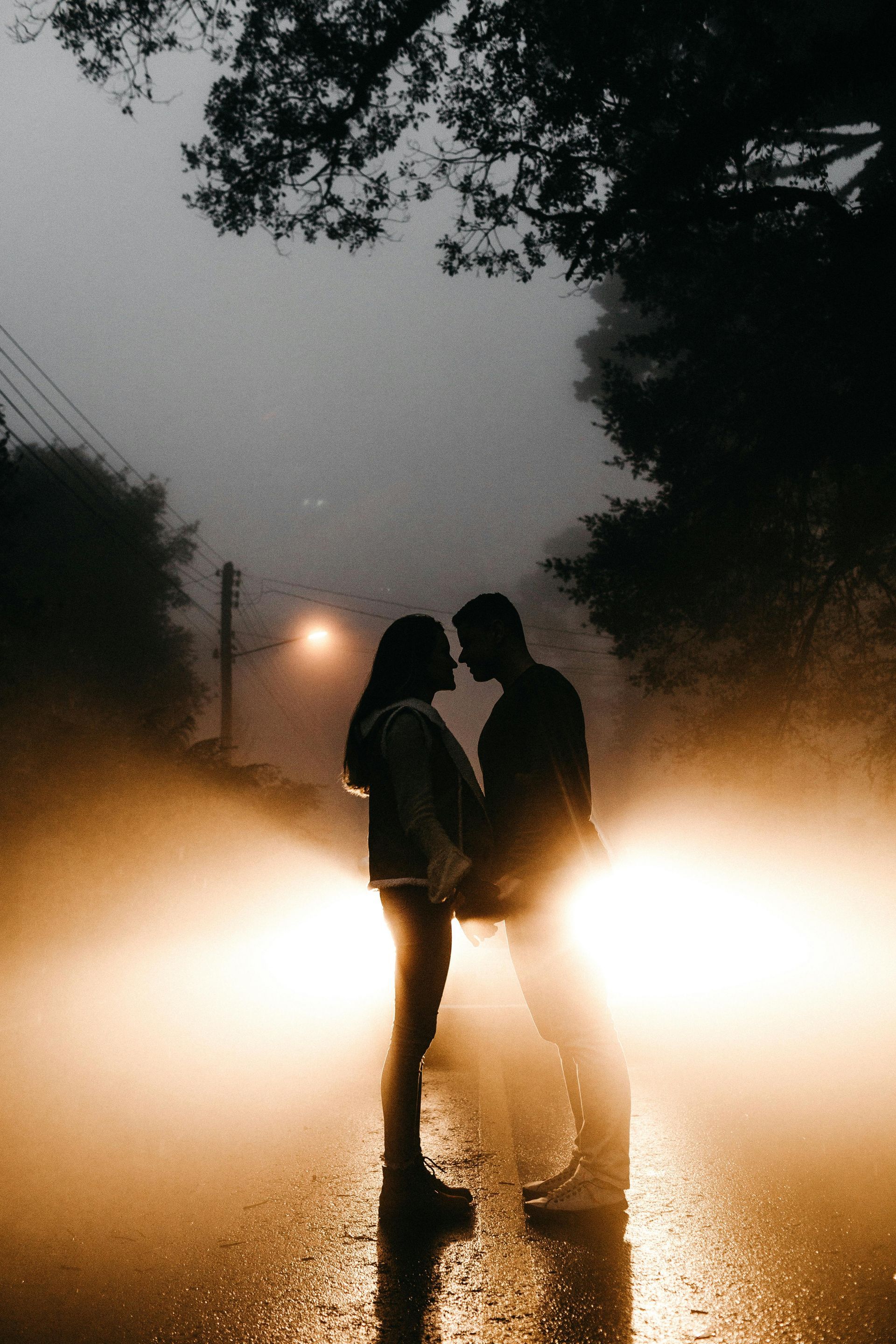 Silhouetted couple facing each other on a dark, rainy road, illuminated from behind by bright vehicle headlights.