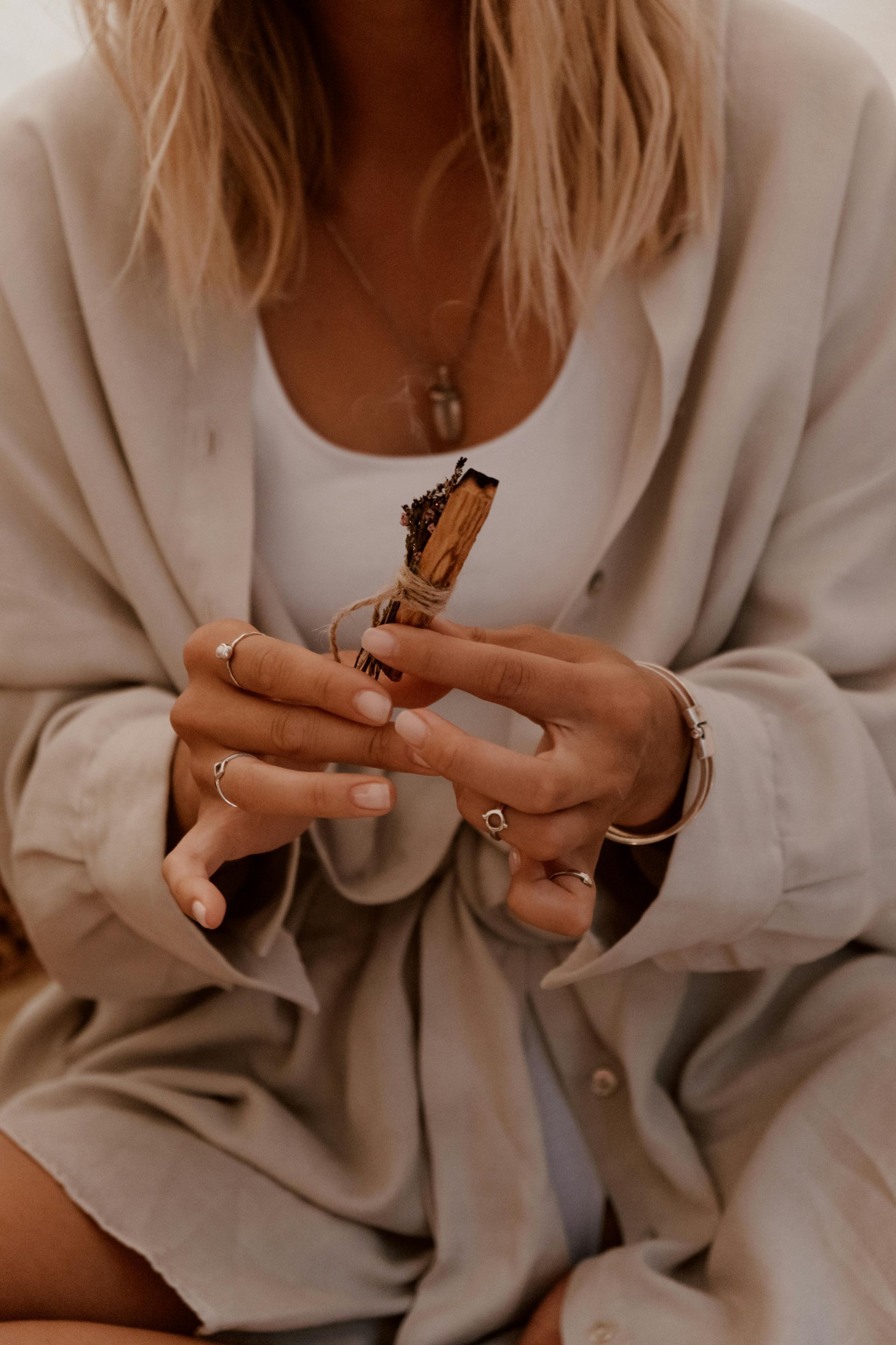 Woman holding a bundle of Palo Santo, wearing a white top and tan linen shirt, hands adorned with rings and bracelets.
