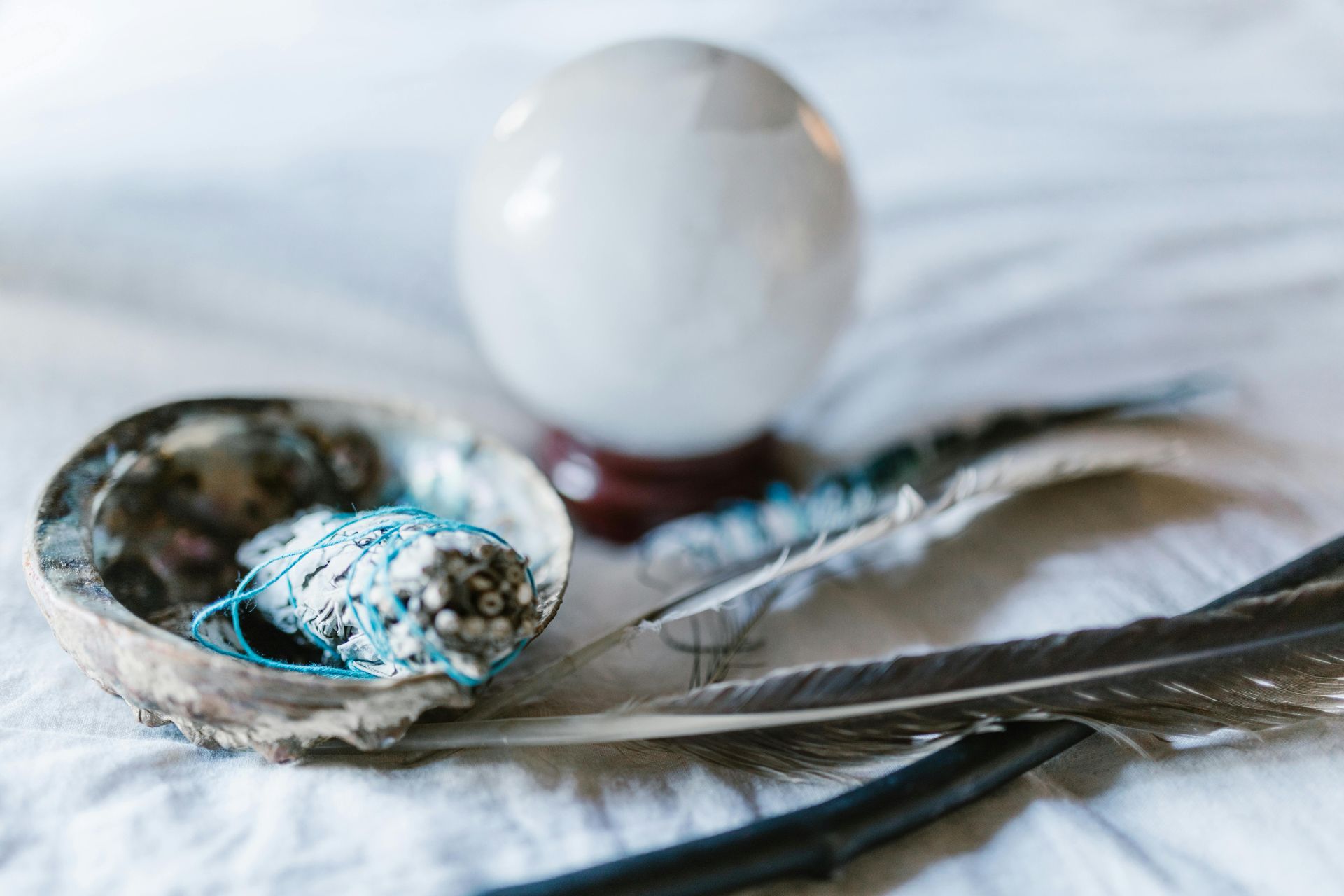 Crystal ball, silver spoon with blue stones, and feather on white fabric.