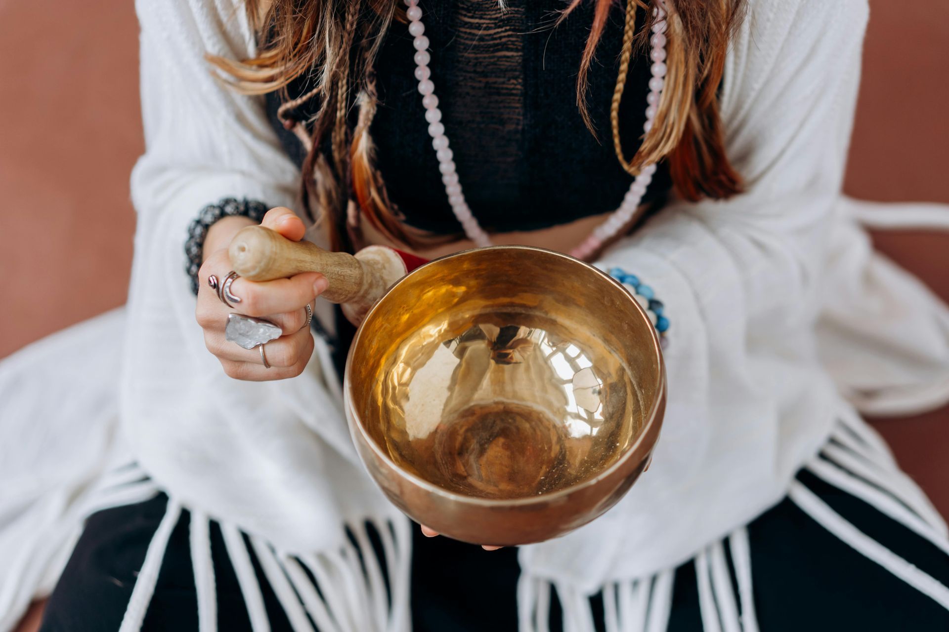 Person seated, holding a golden singing bowl. They are about to play the bowl with a mallet, close up shot.