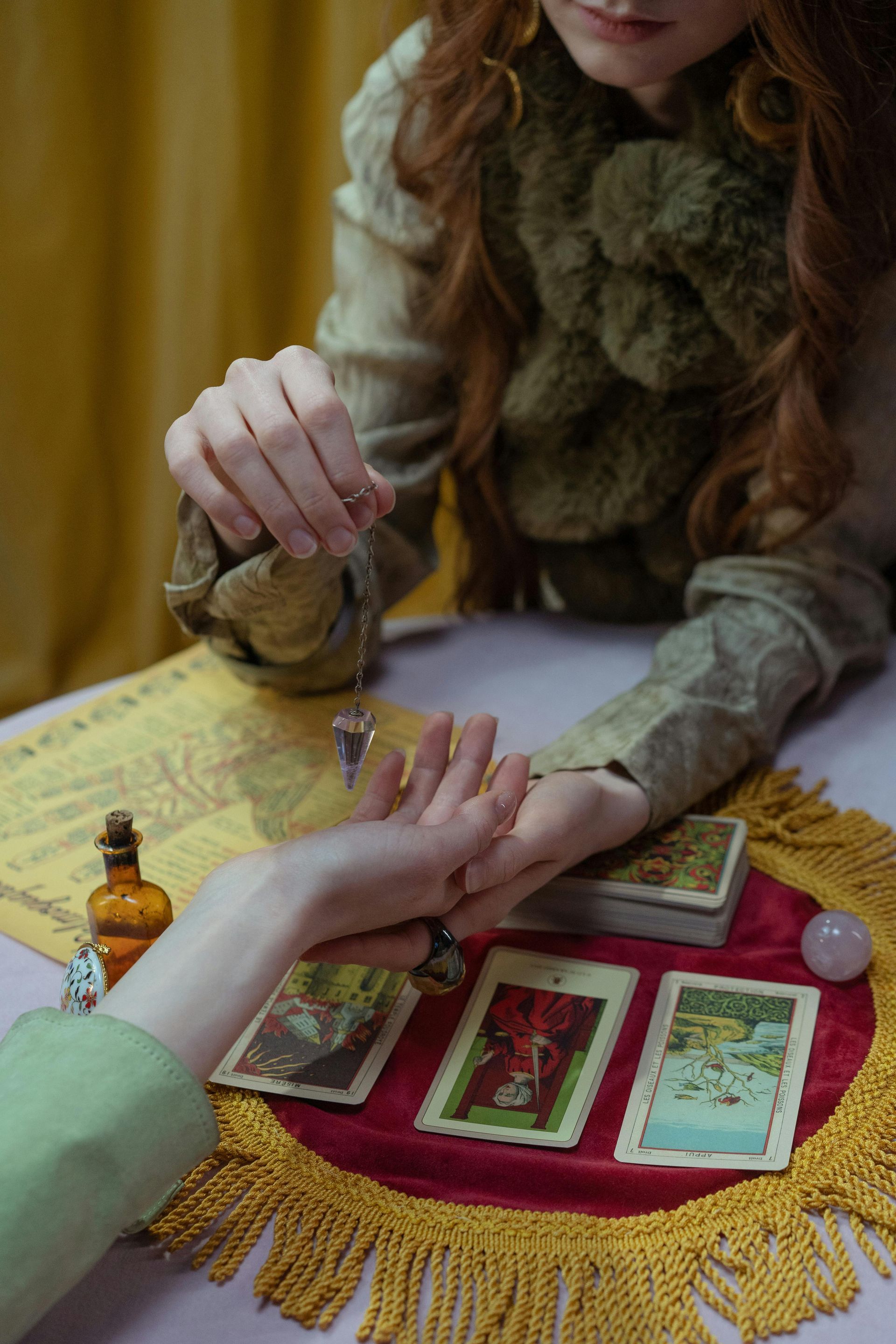 A fortune teller uses a pendulum over a person's hand, surrounded by tarot cards and a crystal ball on a table with a red cloth.