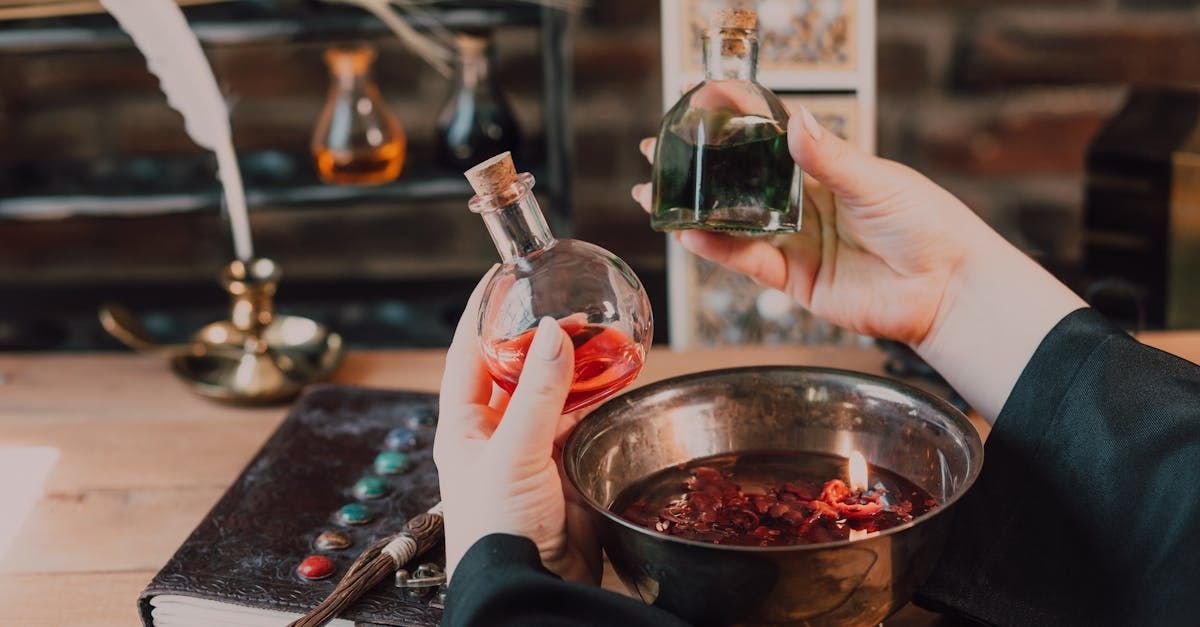 Hands pouring a small vial of dark liquid into a bowl of herbs and red petals, with another potion bottle held nearby.