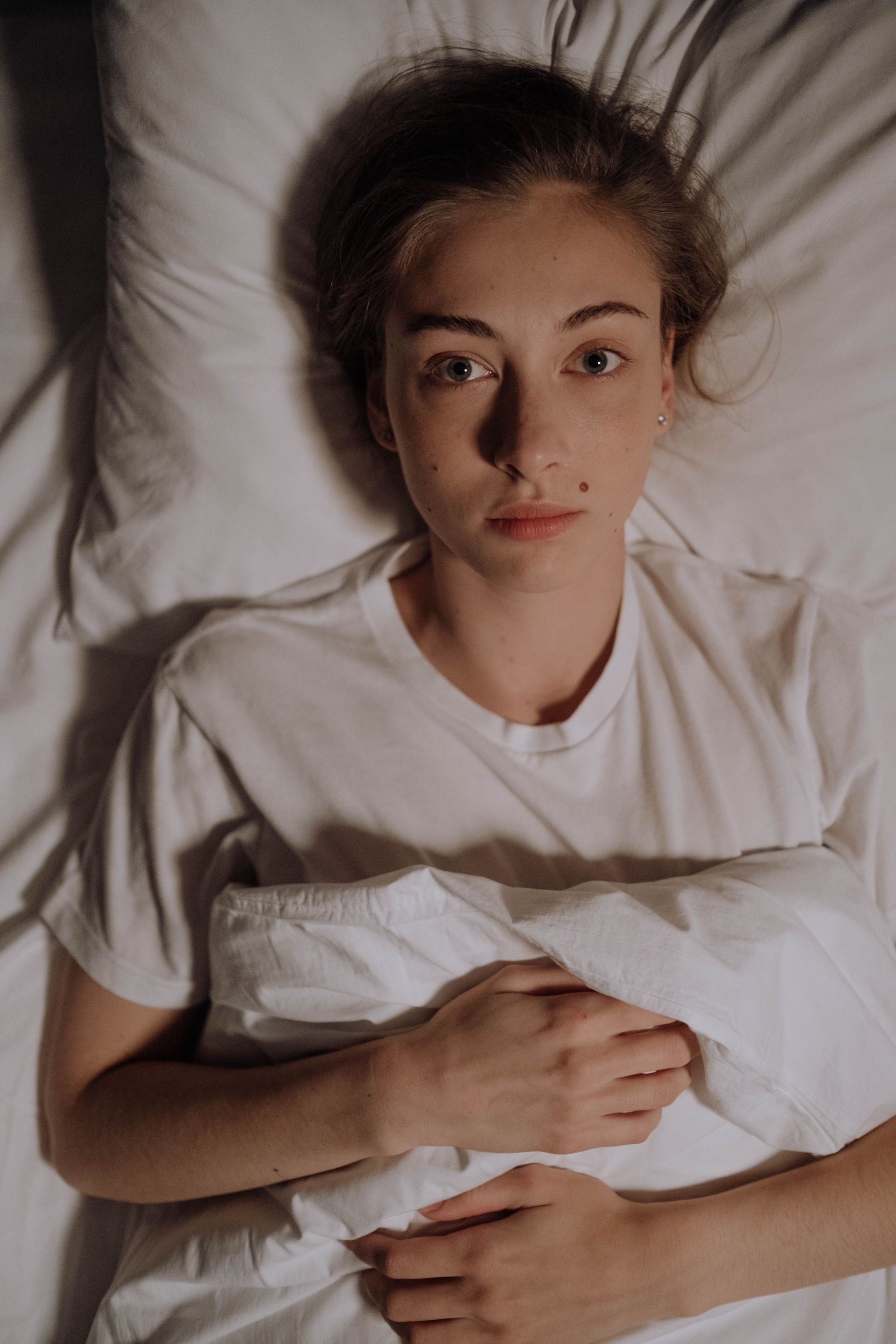 Person lying in bed, holding a white pillow and looking up calmly.