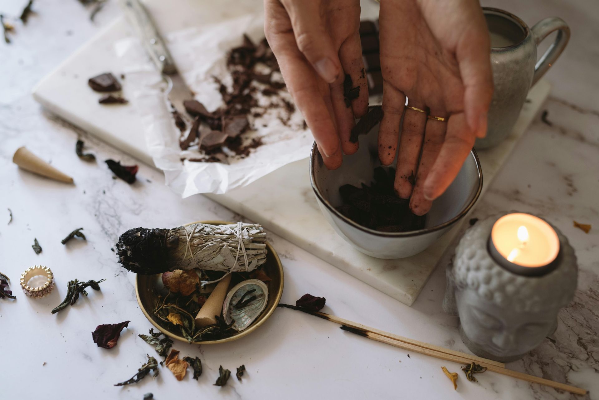 Hands preparing herbs in a bowl, surrounded by candles, smudge stick, and marble surface.