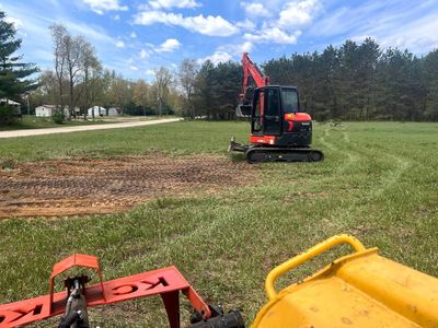 Red excavator working on a grassy field under a partly cloudy sky, with a yellow loader in the foreground.