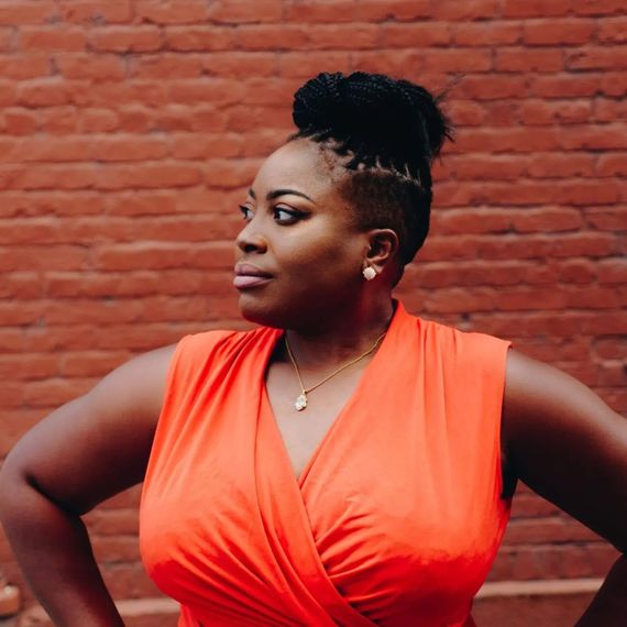 A woman in an orange dress is standing in front of a brick wall.