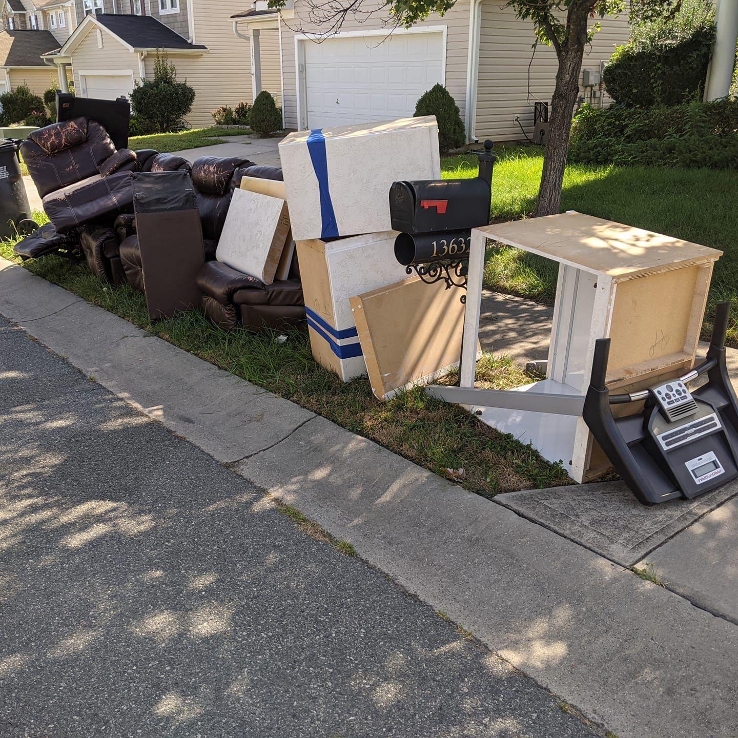 Furniture, a mailbox, and household items piled on a grassy curb for curbside trash collection in front of houses.