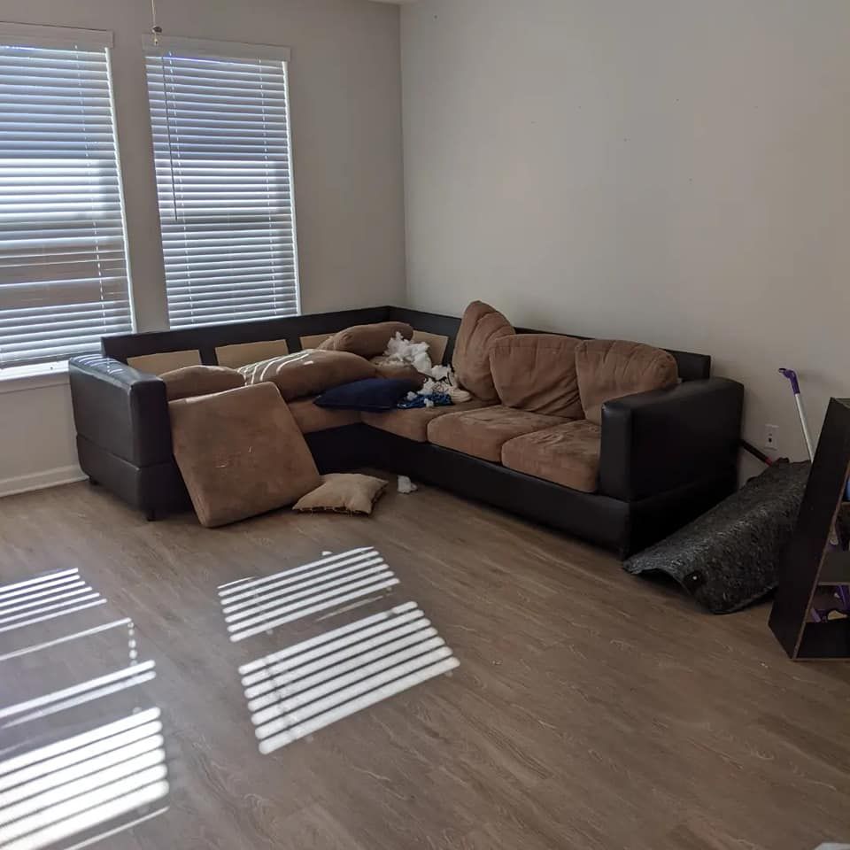 A brown sectional couch sits in the corner of a room with blinds on the windows and wood-look flooring.