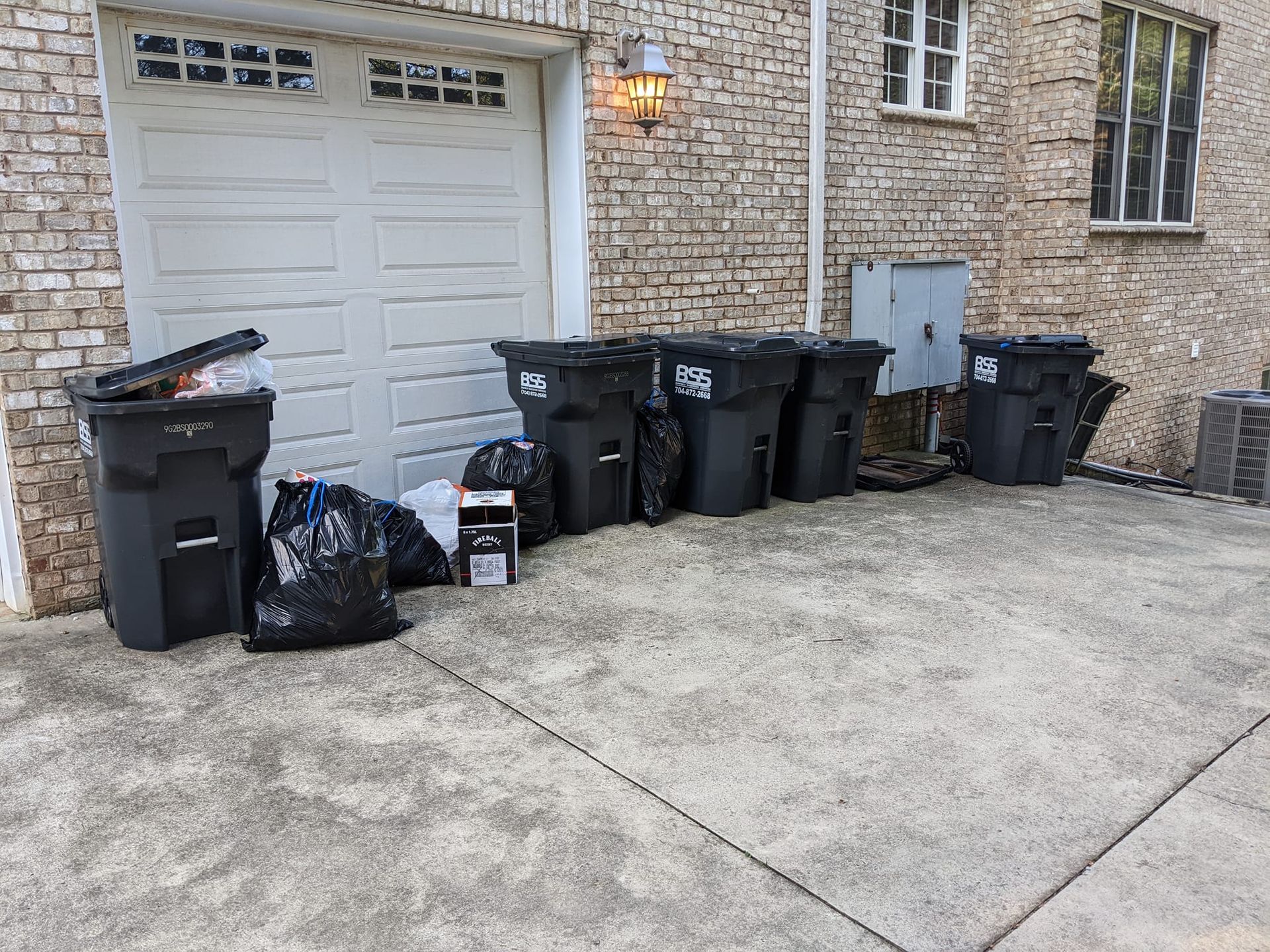 Multiple black trash cans and several filled garbage bags lined up on a concrete driveway against a brick house wall.