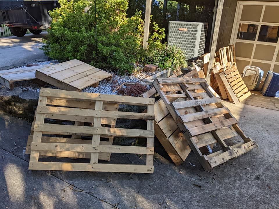 Several wooden pallets are scattered on a paved driveway near a building, bushes, and an outdoor air conditioning unit.