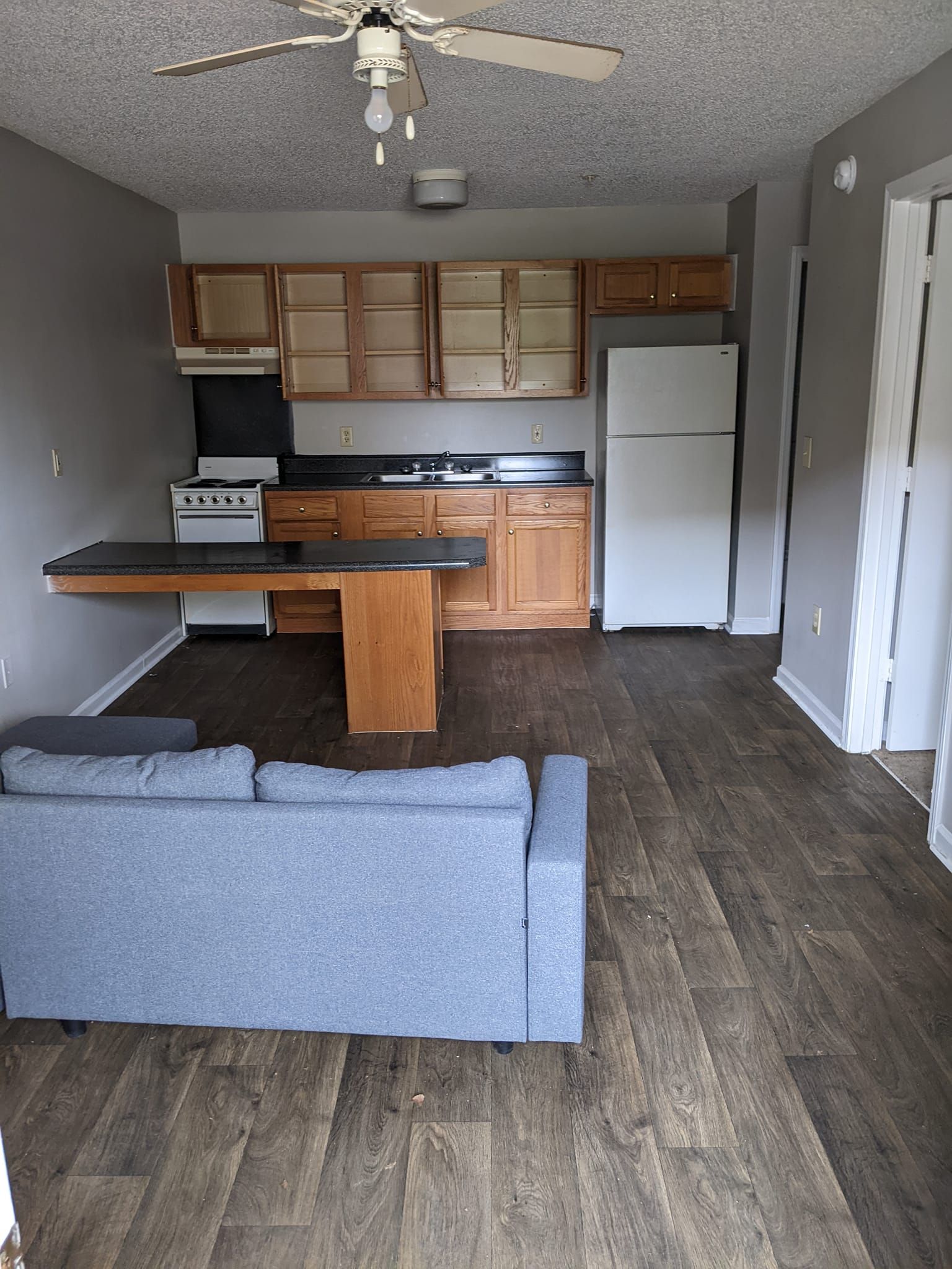 A studio apartment with a gray couch in the foreground, a kitchen with wood cabinets, white appliances, and wood floors.