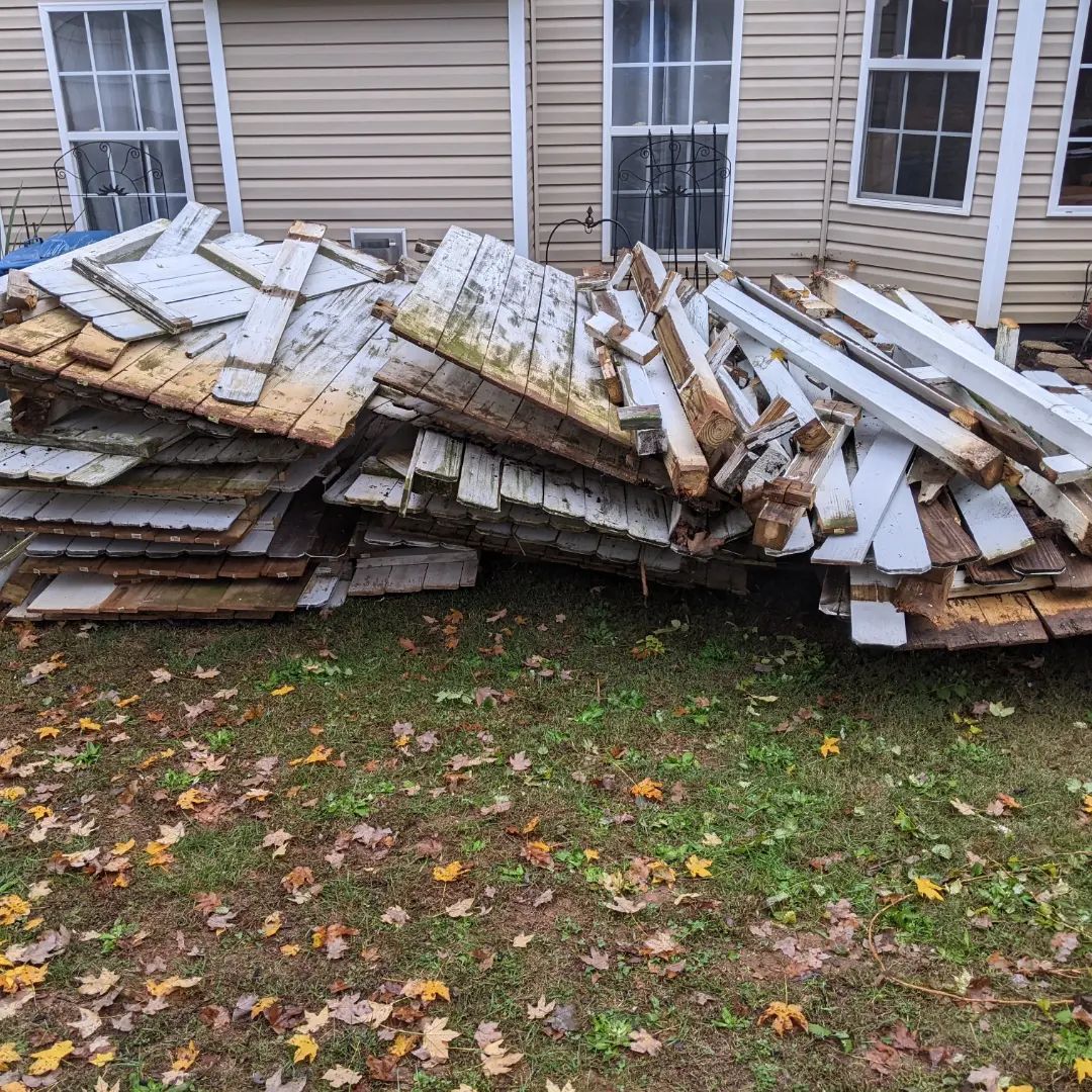 A pile of weathered, white wooden fence panels and debris sits on the grass in front of a house with tan siding.