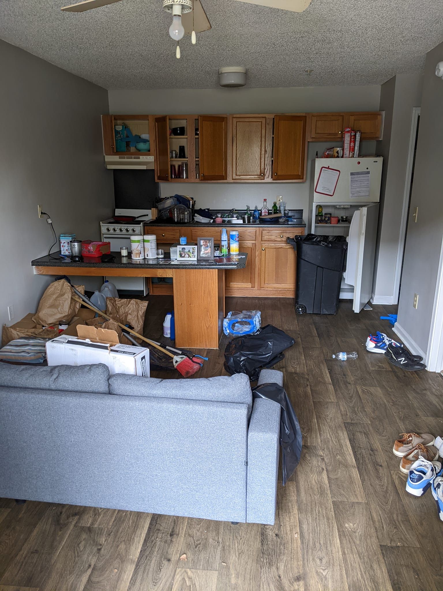 A living area and kitchen with a grey couch, wooden cabinets, a cluttered counter, a refrigerator, and wood-look floors.