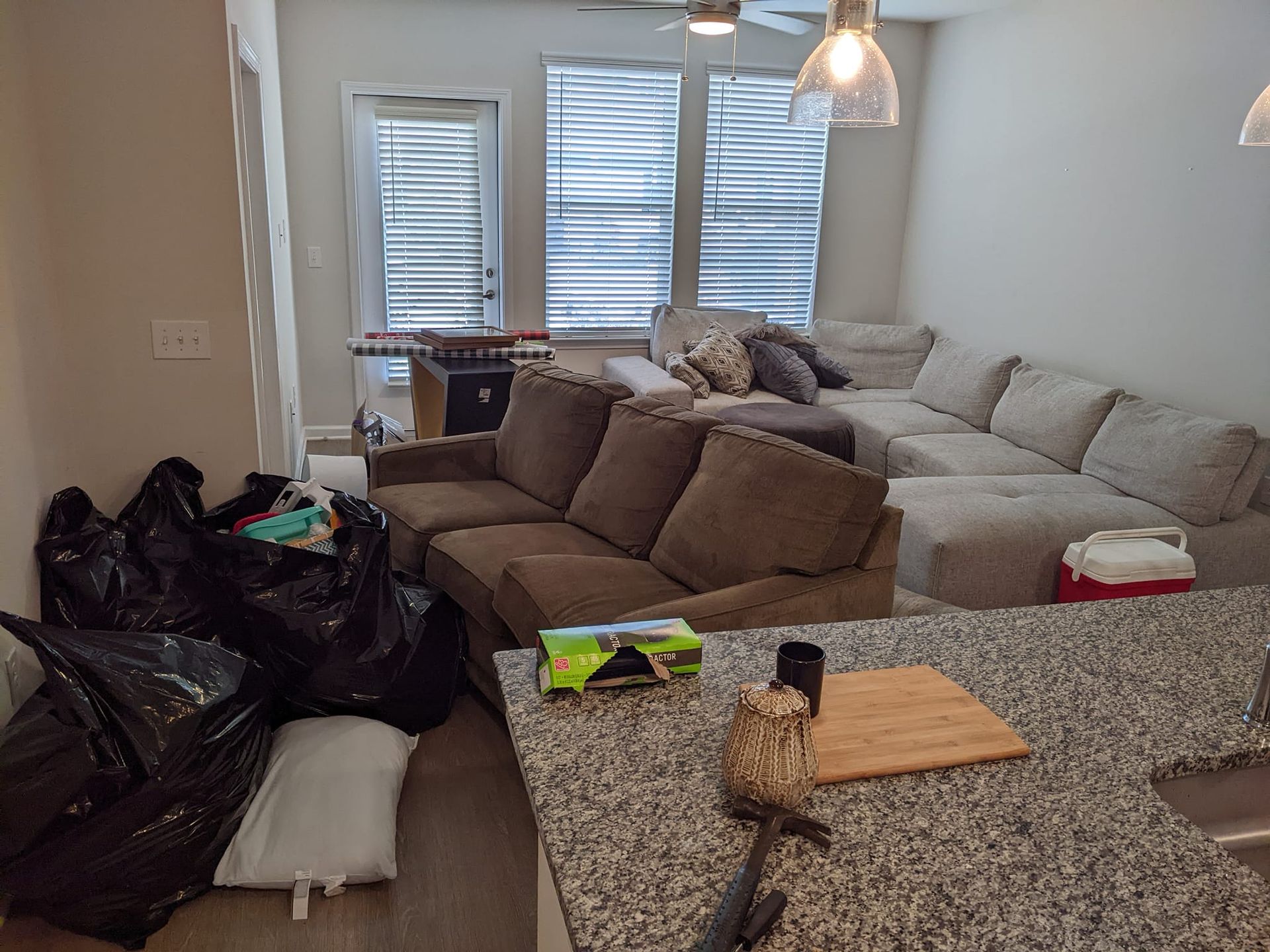 A living room with a large gray sectional sofa, a brown couch, and several trash bags piled on the floor in the foreground.