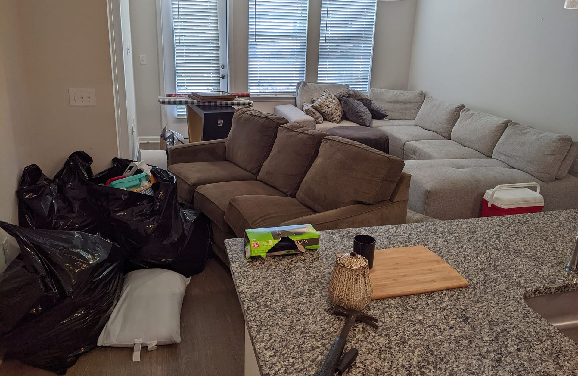 A cluttered living room with a brown sofa, a large grey sectional, and several black trash bags on the floor.