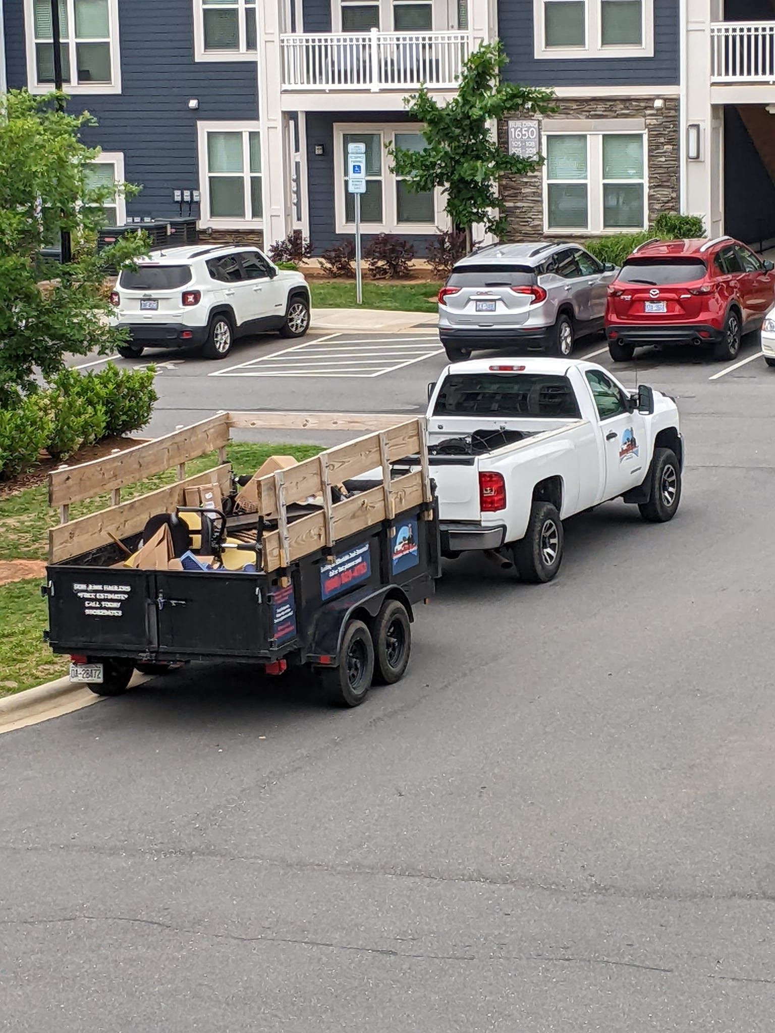 A white pickup truck towing a black utility trailer is parked in front of a residential apartment building.