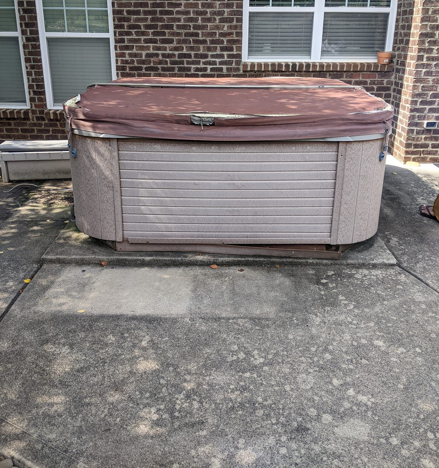 An outdoor hot tub with a faded, worn brown cover sits on a concrete patio in front of a brick house.