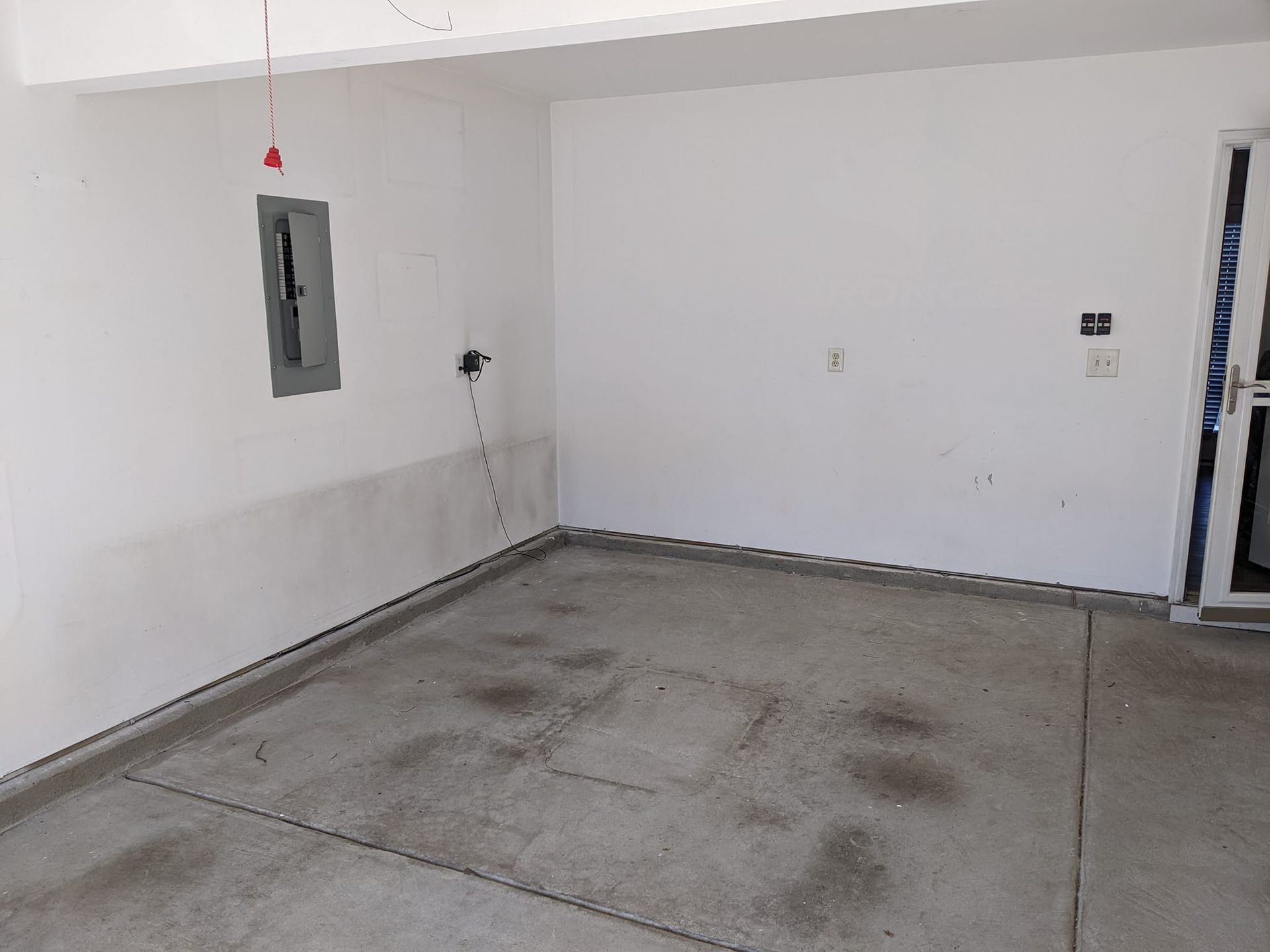 An empty, white-walled garage with a concrete floor, an electrical panel on the left wall, and a door on the right.