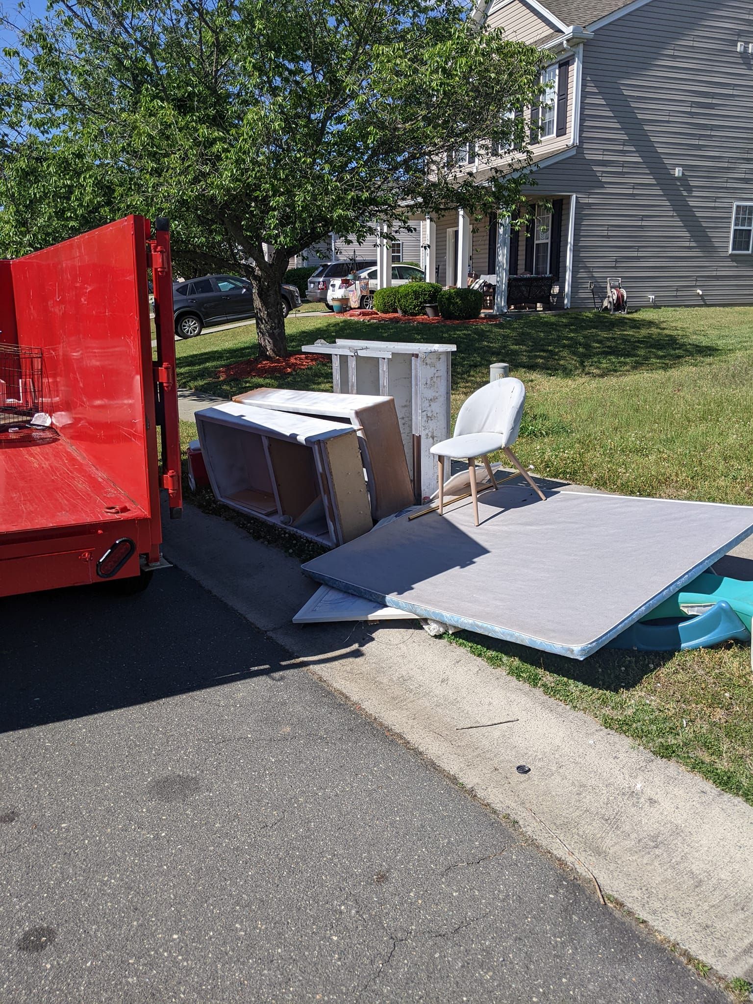 A red dumpster sits on a driveway next to discarded furniture, including a chair and pieces of wood, on a sunny day.