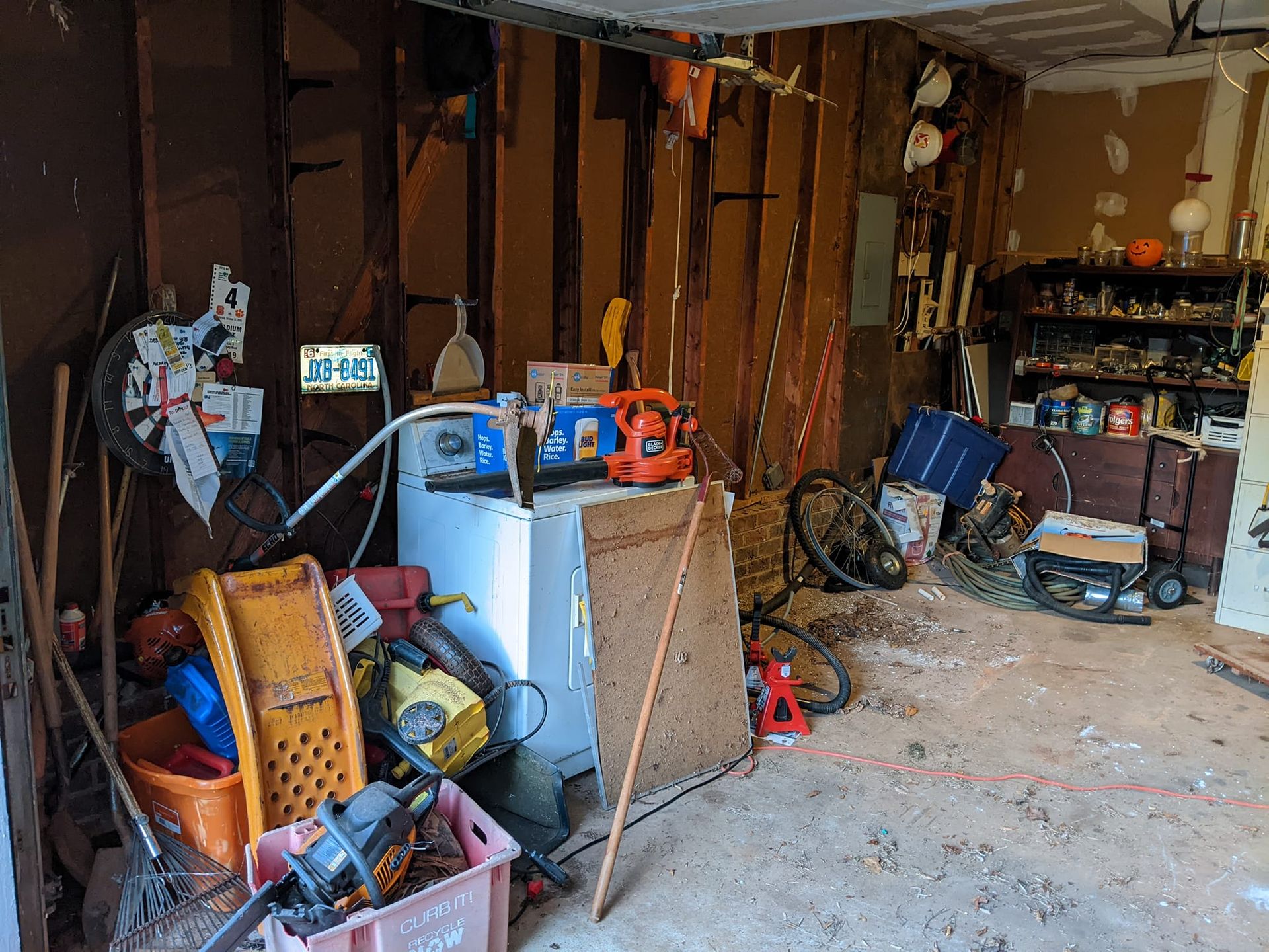 A cluttered garage interior with exposed wall studs, various tools, boxes, a freezer, and scattered household items.