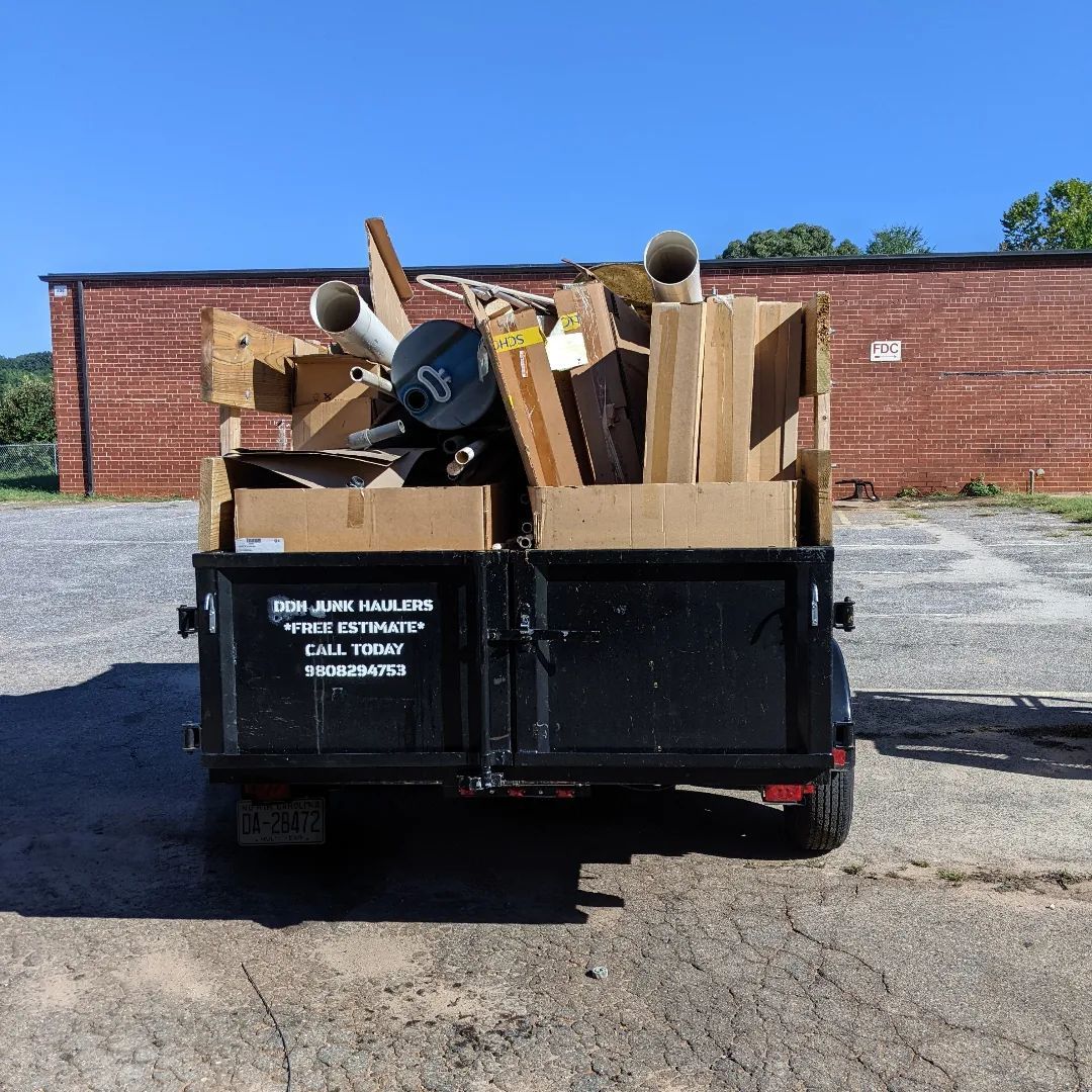 A black utility trailer loaded with cardboard boxes and miscellaneous debris, parked outdoors against a brick wall.