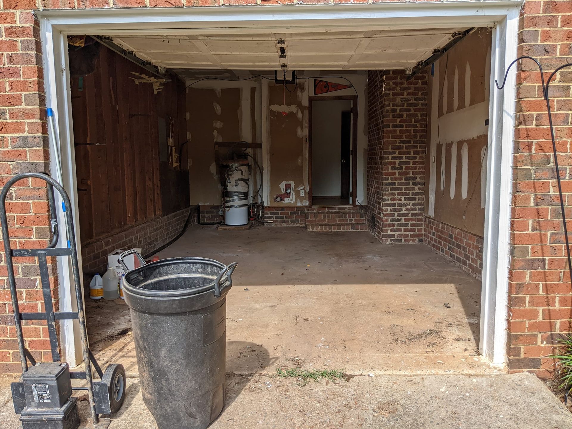 An open residential garage with bare studs on the walls, a concrete floor, a trash can, and a hand truck in the foreground.