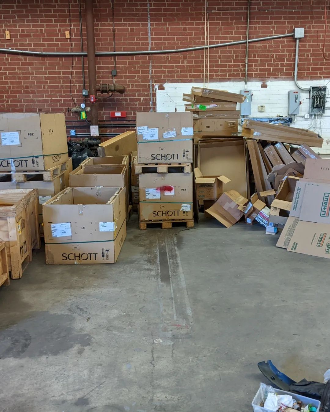 Stacked cardboard boxes and pallets are organized in a warehouse with a brick wall background.