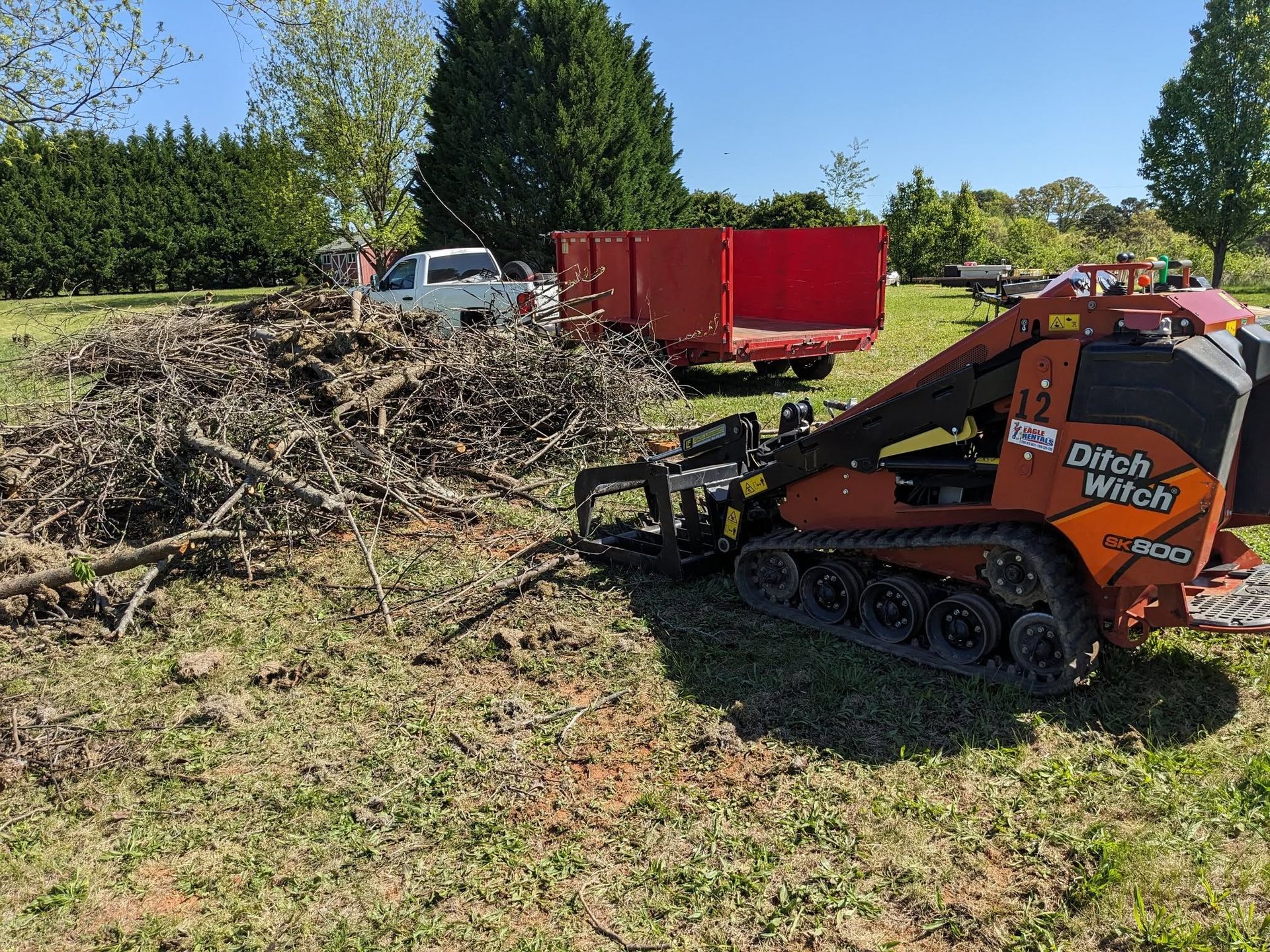 An orange Ditch Witch skid steer with a grapple attachment sits in a field next to a large brush pile and red dump trailer.