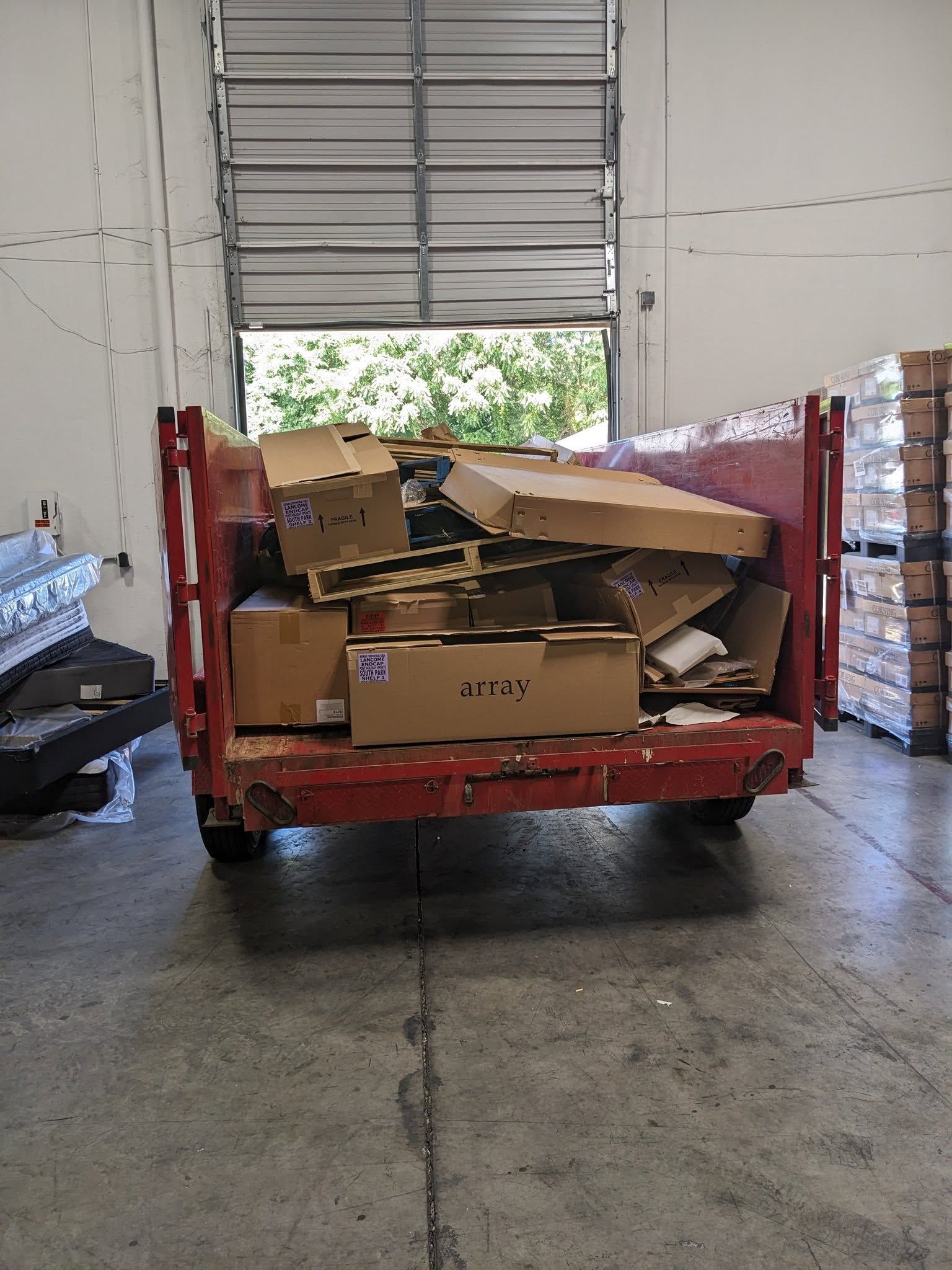 A red dumpster filled with various cardboard boxes parked inside a warehouse with a large roll-up door in the background.