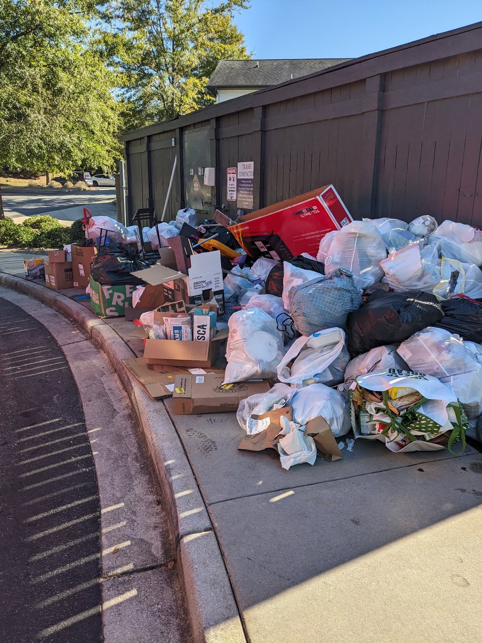 An overflowing pile of trash bags and cardboard boxes stacked against a brown fence on a paved sidewalk.