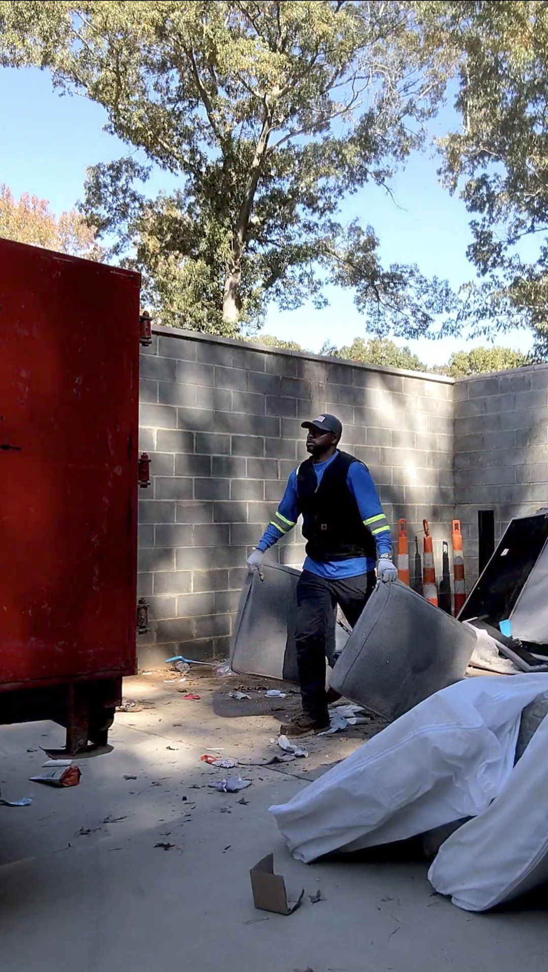 A worker in a high-visibility vest moves a couch cushion near a dumpster in an outdoor construction area.