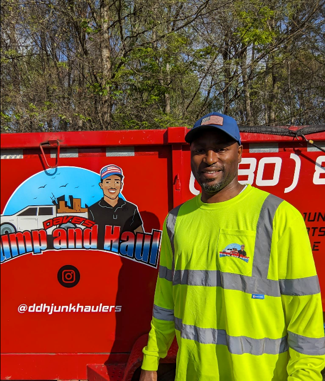 Man in neon safety shirt and blue cap standing by a red hauling truck with a cartoon logo.