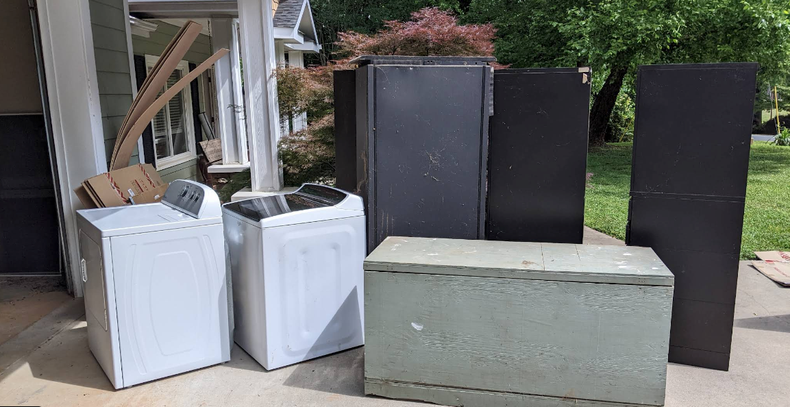 Assorted appliances and cabinets stacked outside next to a house porch, including white and black units and a gray block.
