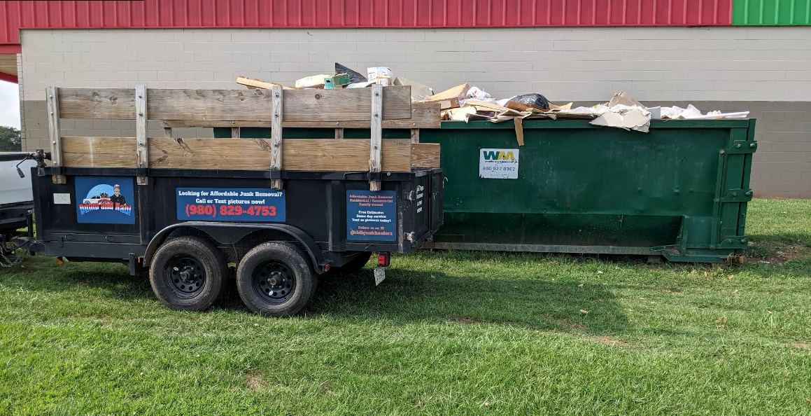 Blue dump trailer beside a green dumpster on grass in front of a red building.