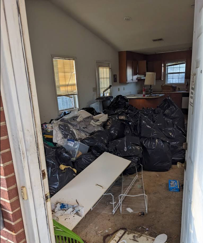 Cluttered kitchen and living area filled with trash bags, debris, and a broken table.